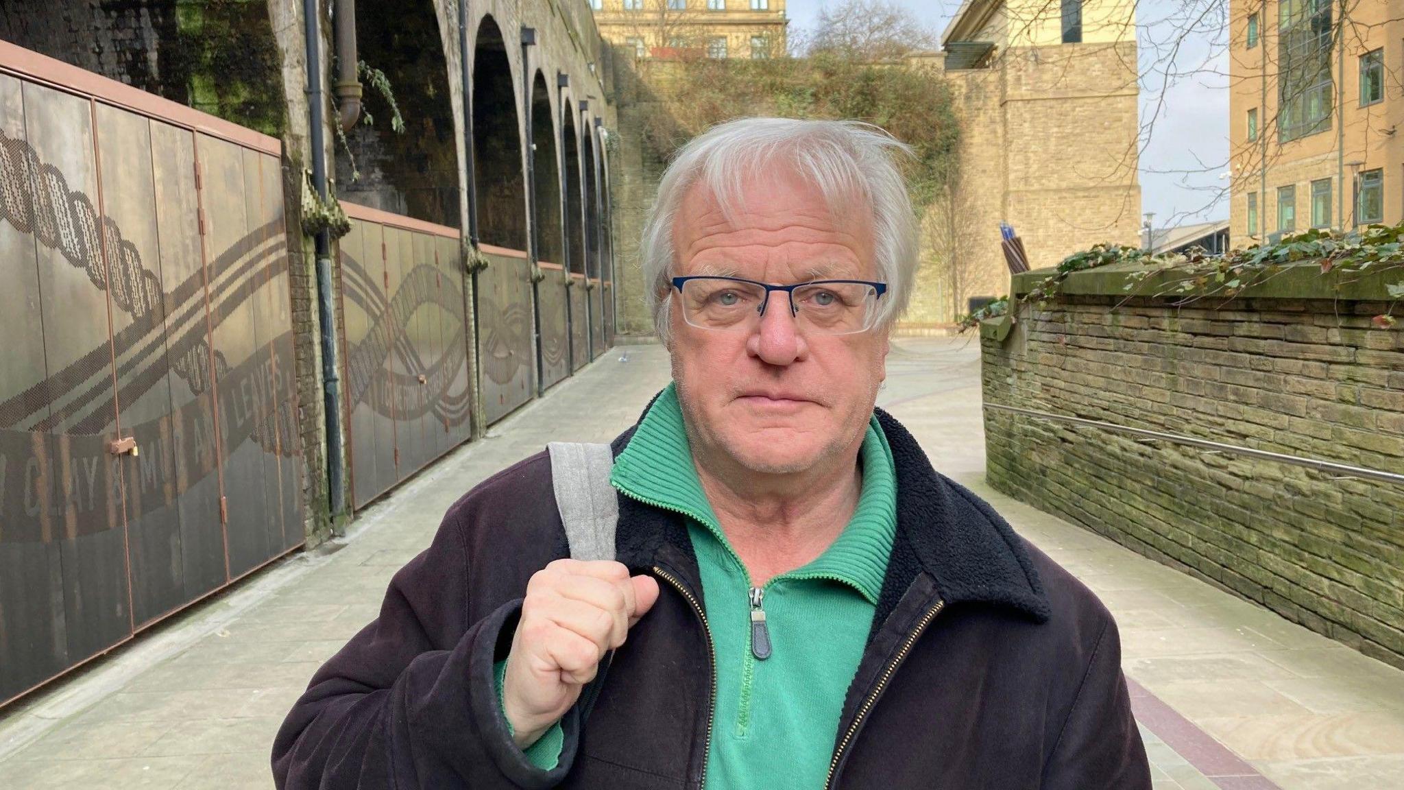 A man with white hair and glasses wearing a green zip-up top and black coat. 