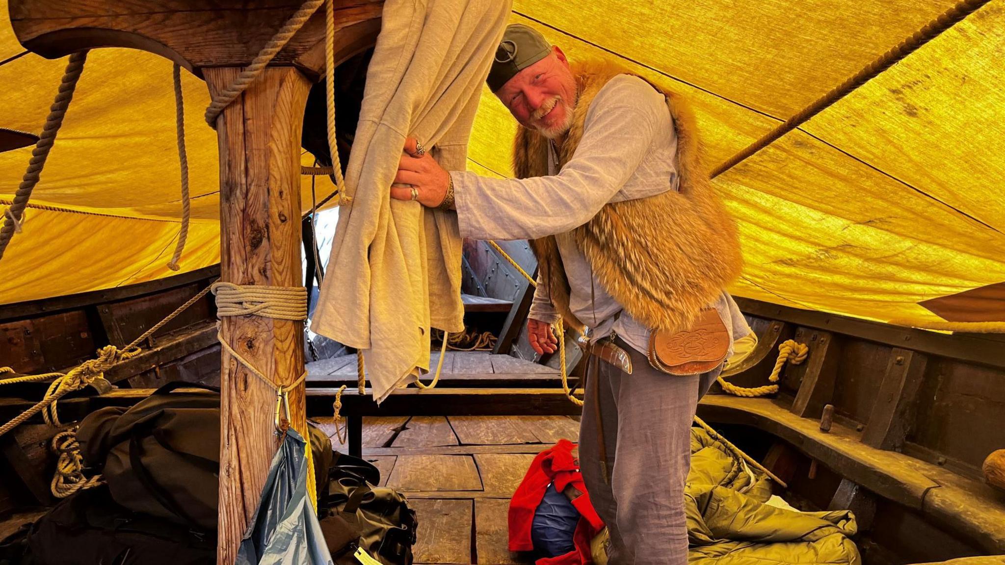 A man is stood, slightly hunched, under a large yellow canopy on a wooden boat. He is looking at the camera and smiling. He has a grey goatee beard and is wearing a Viking-style helmet, a light grey long-sleeve top, grey jeans and a fluffy brown vest. He is clutching some white sheets next to a wooden pillar in the middle of the ship.