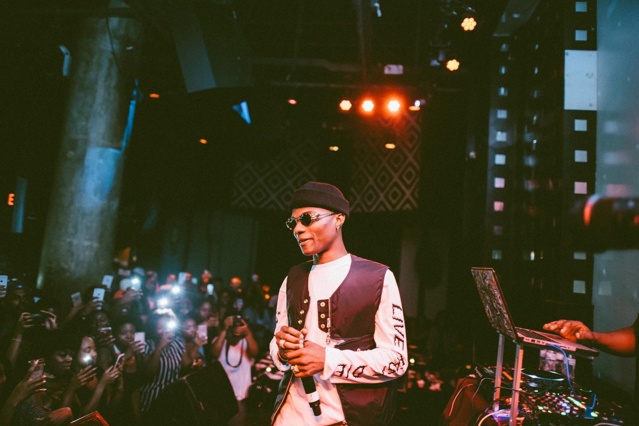 Wizkid in round sunglass and a brown beanie, cream sweatshirt and brown waistcoat, holds a microphone on a small stage. To his right can be seen a small mixing desk and to left a group of mainly female fans taking photos of him with their phones.