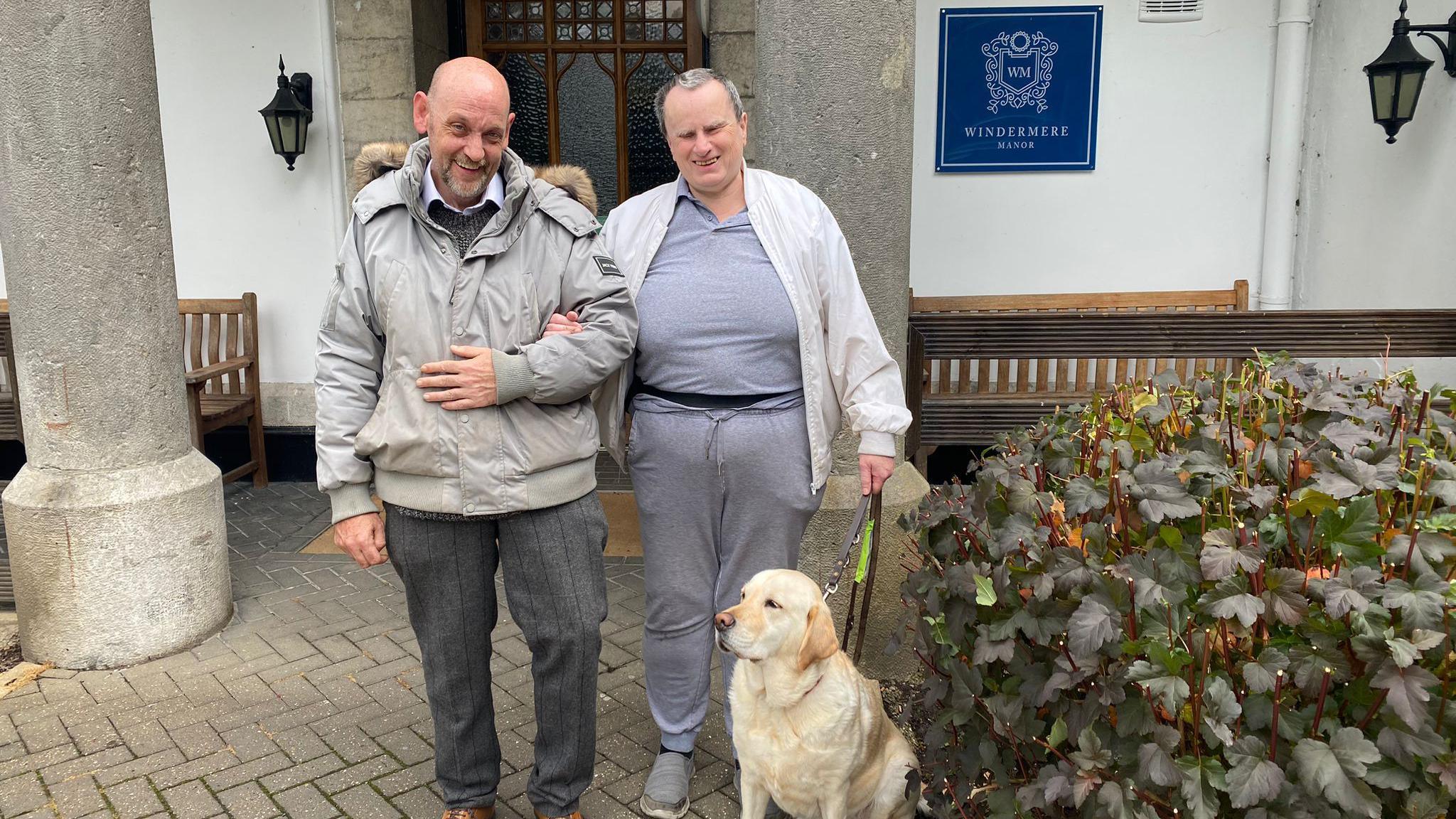 Gareth and Lee outside the Windermere Manor Hotel. They are linking arms with the each other and Lee's guide dog is sitting in front of them 