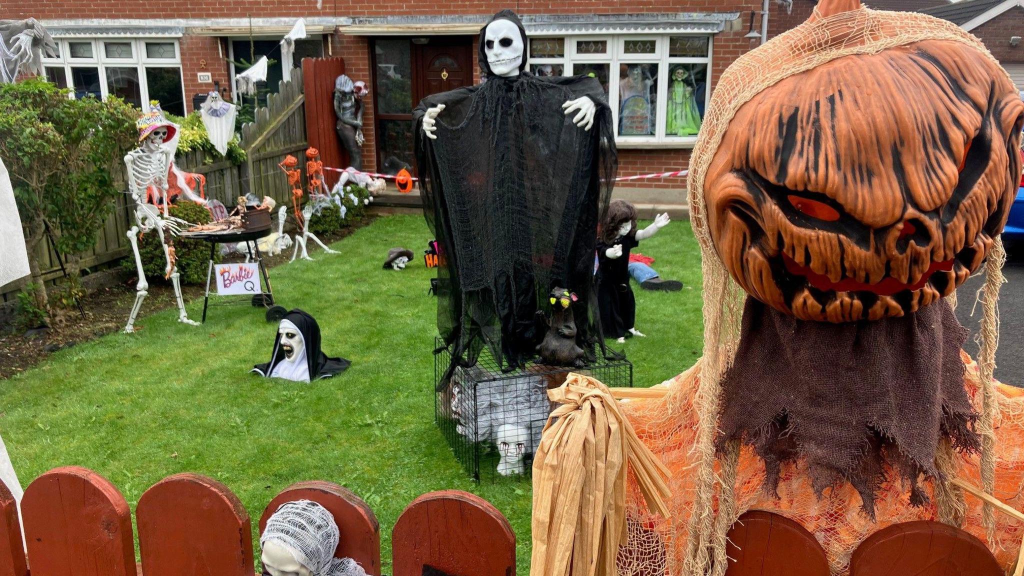 A brick house with Halloween decorations. There's an orange pumpkin headed monster at the fence. A nun buried in the grass, a skeleton at the barbecue. A skull headed monster is standing on top a cage.