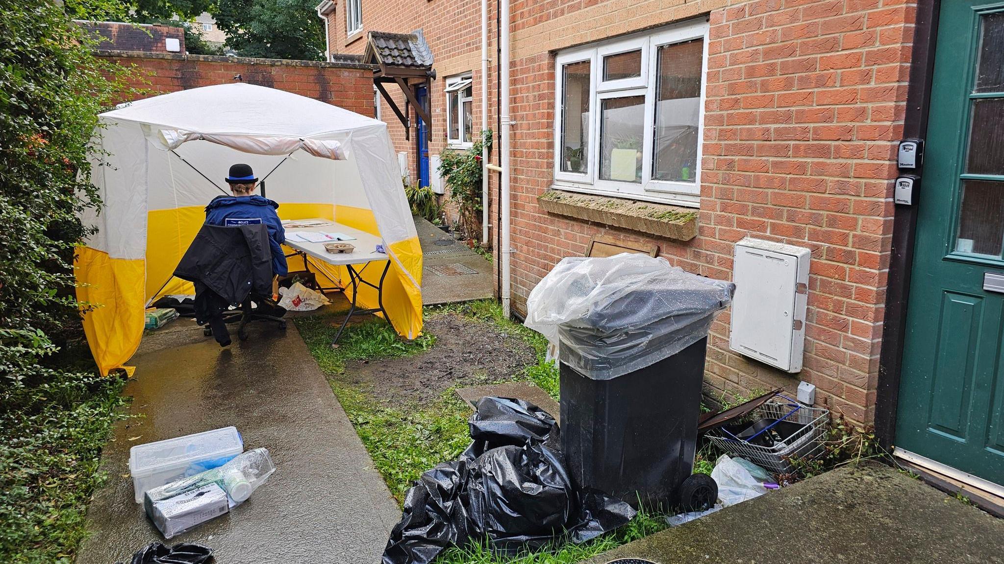 A police forensic tent set up outside a home in Juniper Close where police believe Mr Wheeler was attacked. The house is made of red bricks and there is a concrete walkway leading to the door. There is a female police officer sitting at a desk inside the tent. It is raining and there are overflowing bins outside the front door. 