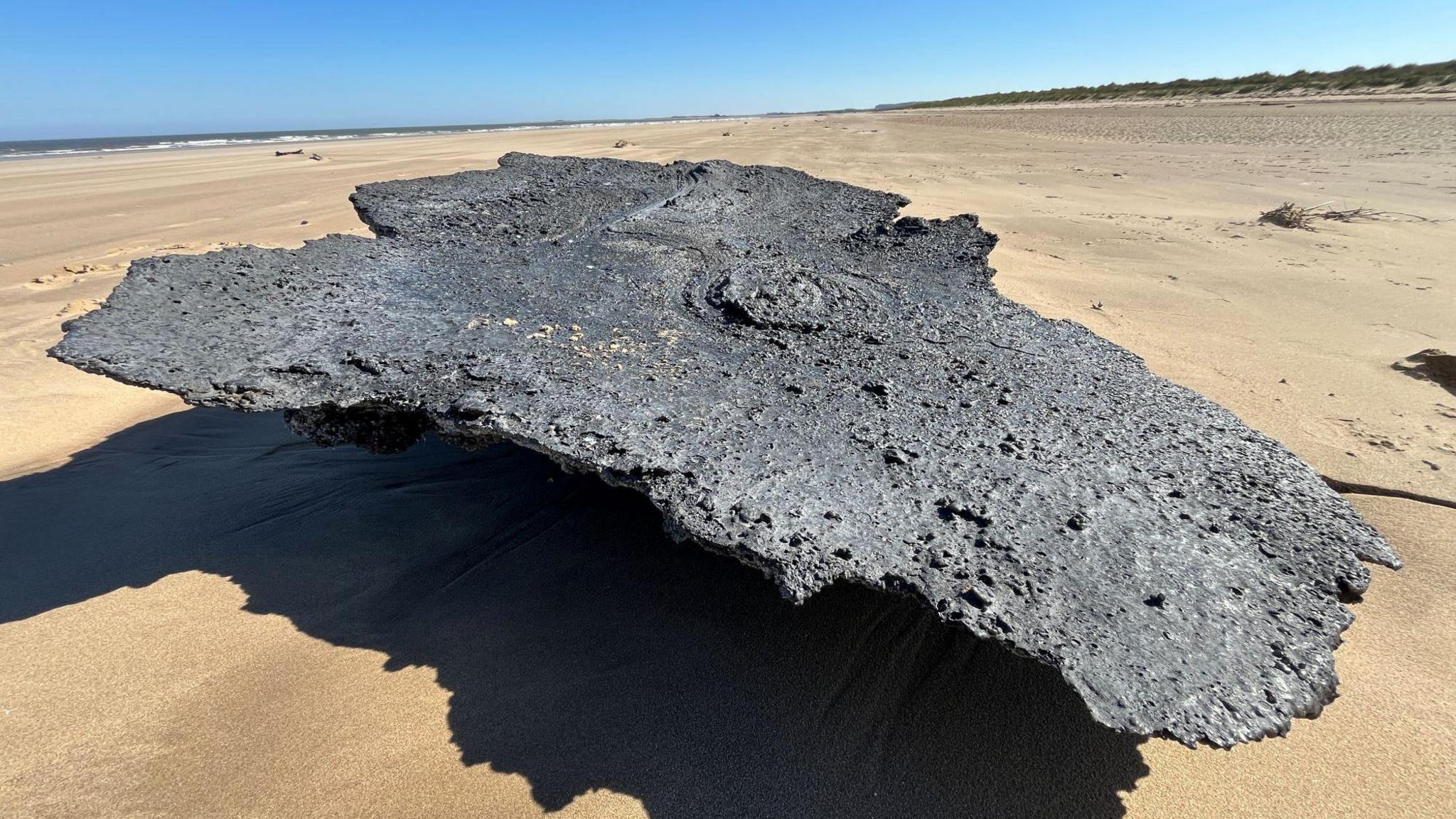 A large clump of charred plastic sat on a golden sandy beach. The sky is blue. Sand dunes are to the right of the plastic and the sea to the left.