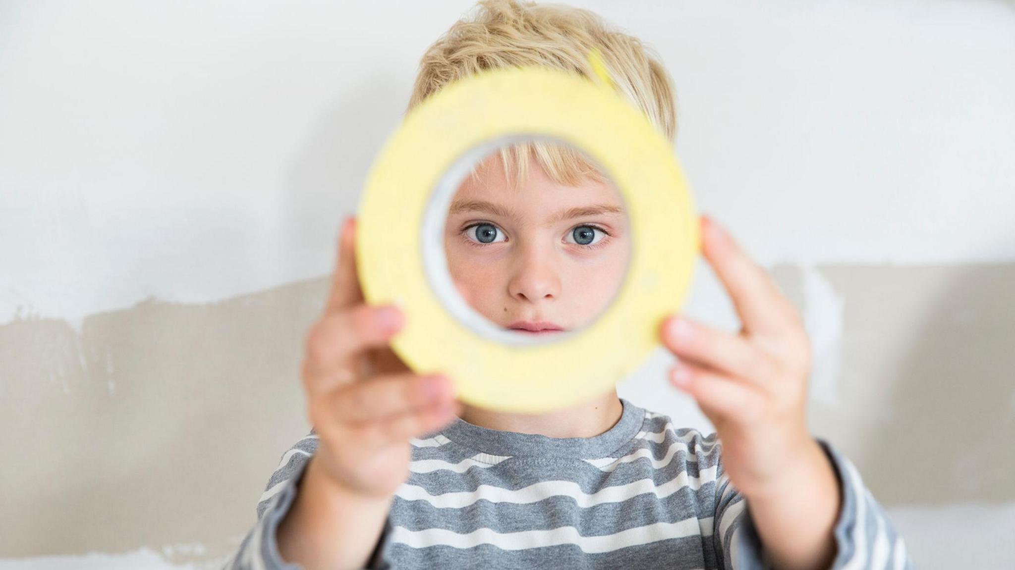 A boy stands holding a roll of tape and peering through it. He wears a grey and white stripy top and has blonde hair.