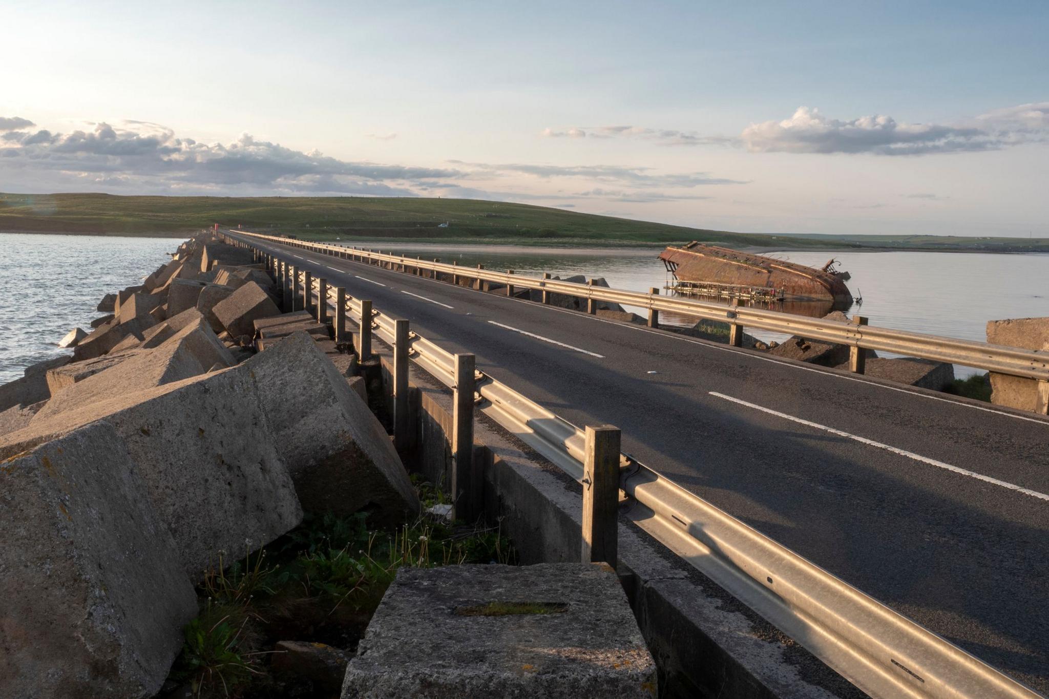 A road across a causeway, with water on either side and shipwrecked boat on the right hand side of the road.