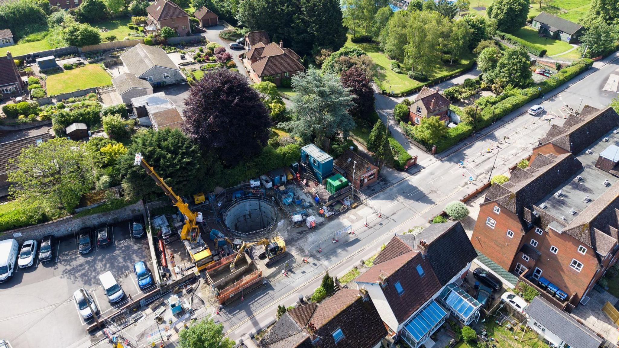 Another overhead drone shot, but higher above, showing the deep hole that is the tank in the centre. The tank is surrounded by the construction site, the partially closed Weymouth Road next to it and housing.