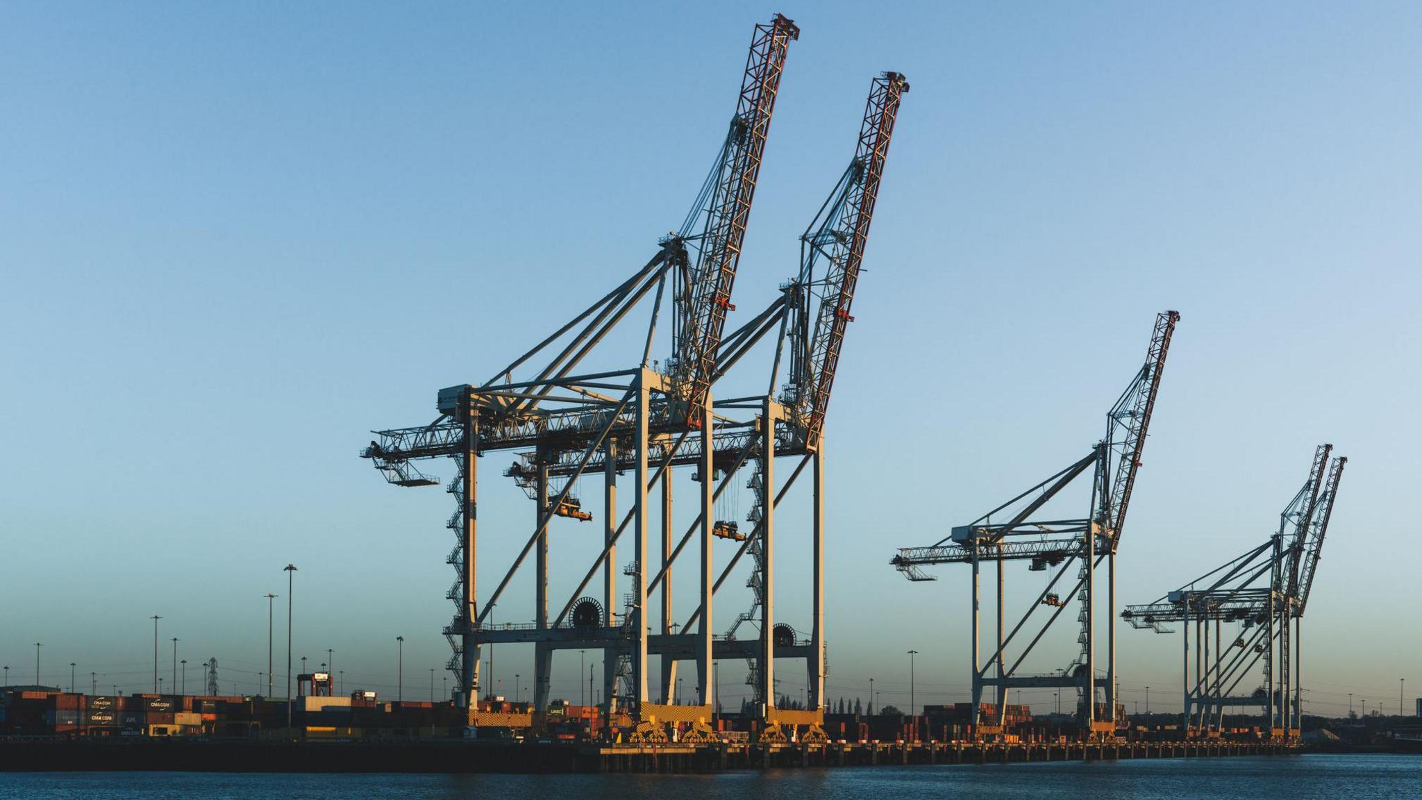 Five enormous container cranes sit on the docks next to the water. The sky behind is completely blue.