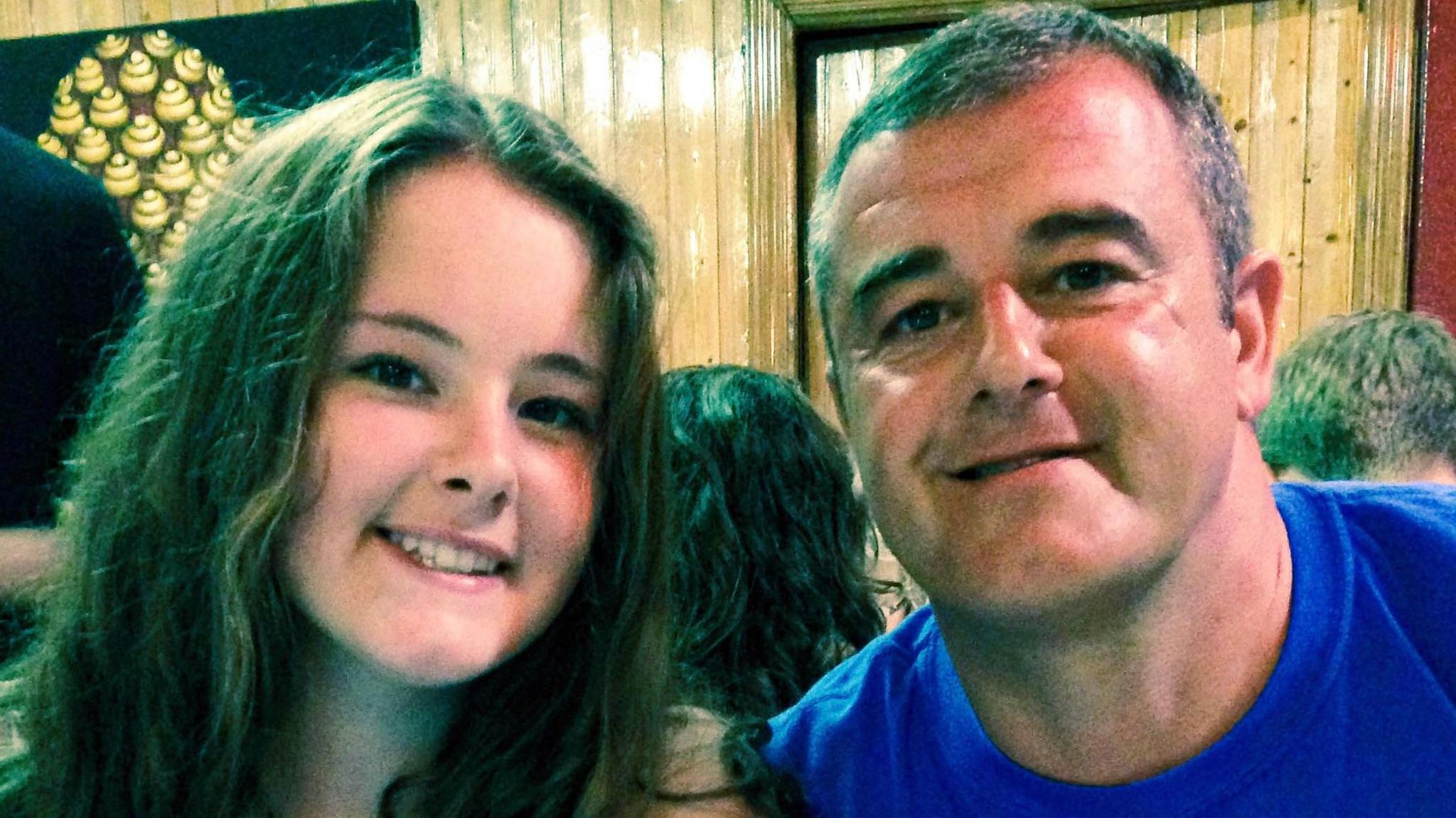 A family photo of Emily and her father David. They are sitting close together indoors, smiling and posing for a photo. Emily, on the left, has long brown hair, and David, on the right has short greyish hair and is wearing a blue shirt. Behind them is wooden panelling.
