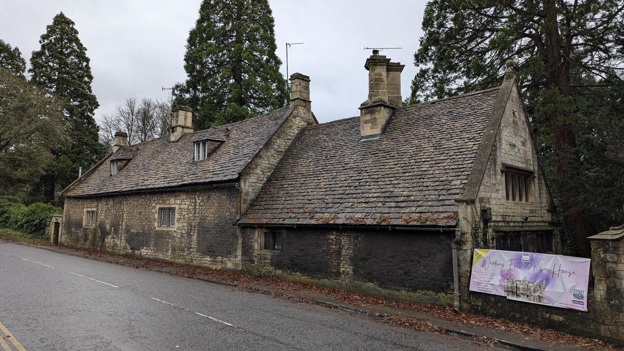 The two stone cottages, viewed from the road. They are covered with large dark marks on the stone, and have small windows set into the walls and pointed roofs. 