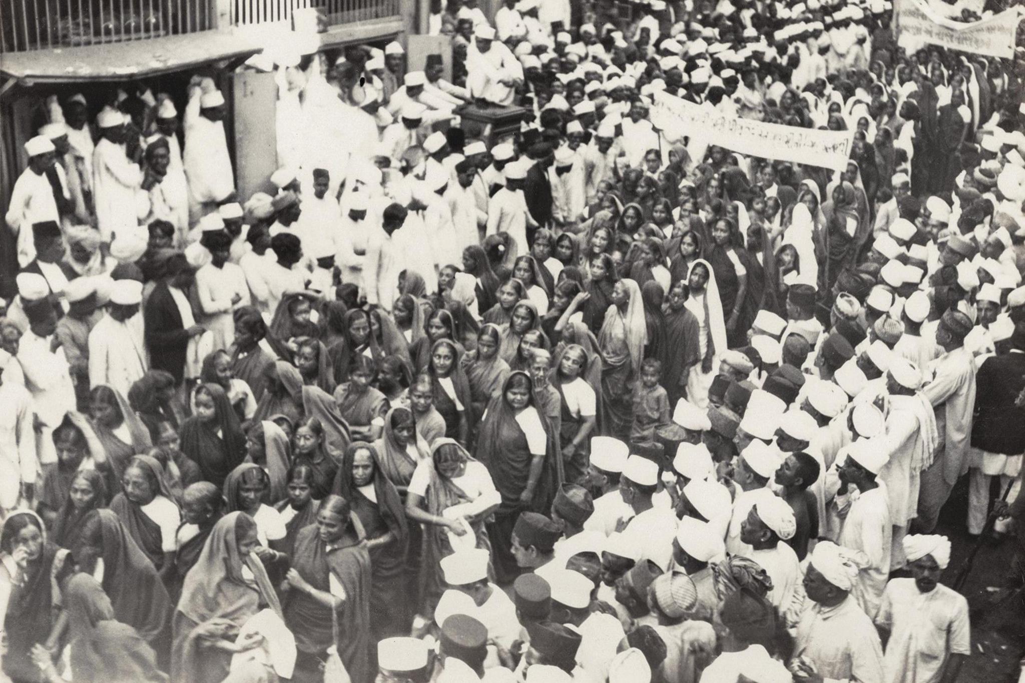 A procession led by women to encourage hand spinning in Mumbai passes through a busy street in India. The women are all wearing saris and have their heads covered. They are surrounded by men, most of whom are wearing caps and kurtas, on both sides of the street.