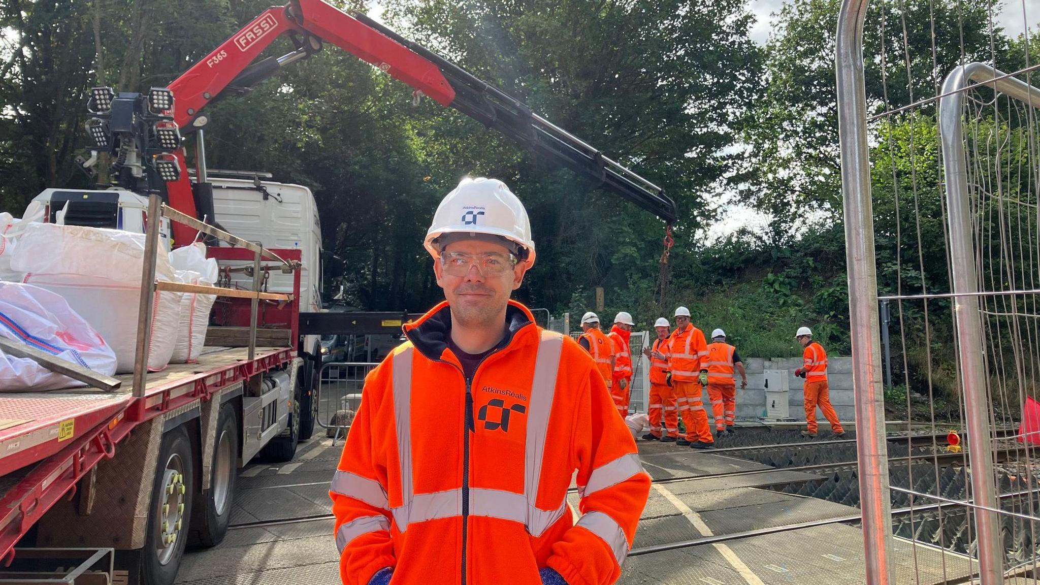 A man stands in front of a group of workers on a railway line, all wearing orange hi-vis clothing and white hard hats. He is looking at the camera and smiling, and has his hands in his jacket pockets. There is also a large lorry seen on the level crossing in the background.