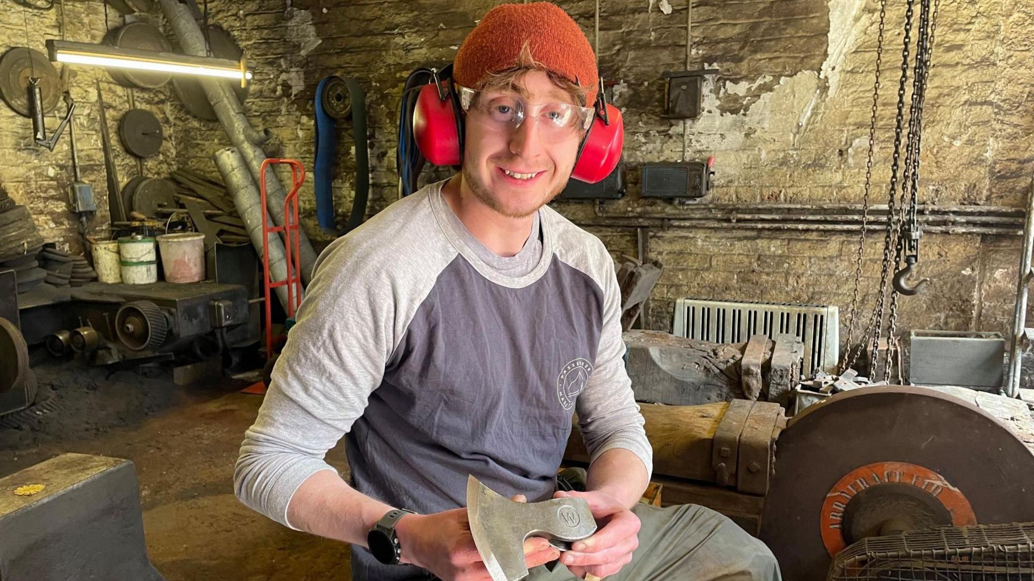 A man with an orange beanie hat and red ear defenders holds up an axe head