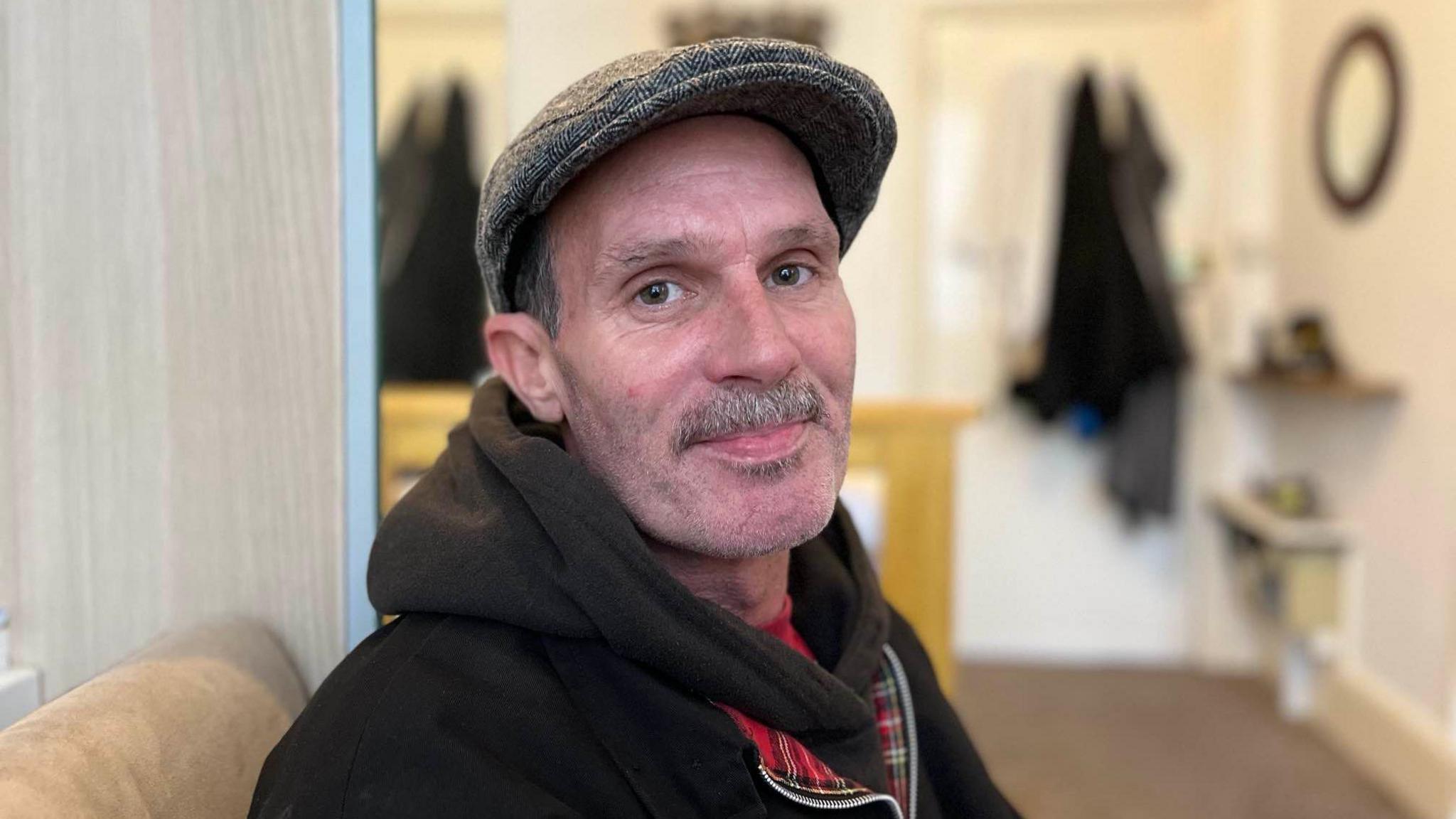 Close up of Steve Hogg inside his home in Hove. He is wearing a tweed cap, dark coat and scarf and has a greying moustache.