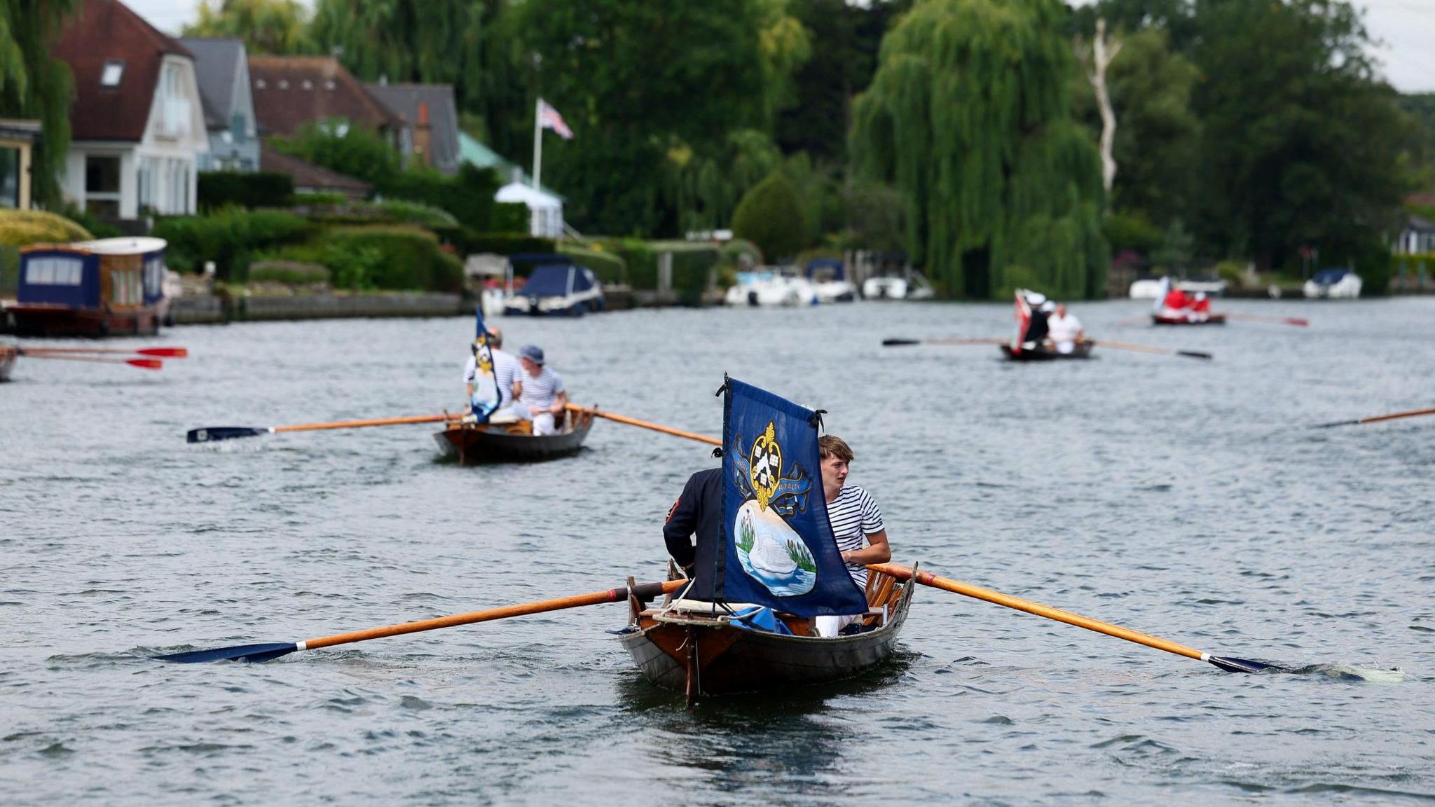A group of traditional wooden rowing boats on a river, each crewed by individuals dressed in nautical uniform. The closest boat prominently displays a blue flag with a swan and a crown on it. The river is lined with riverside homes, boats, and greenery.