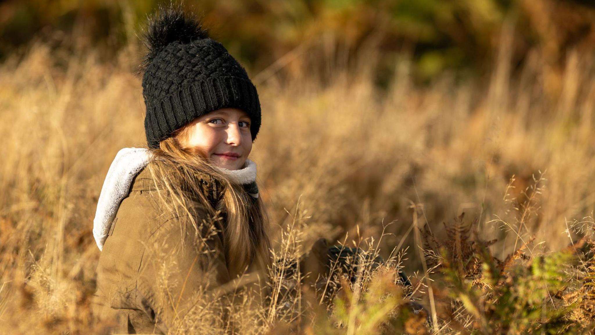 Photograph of 10-year-old Jamie, among the yellow grass. She wears a parka jacket with a fur trim, and wears a black bobble hat. She has long brown hair and blue eyes. There is an orange haze over the image.