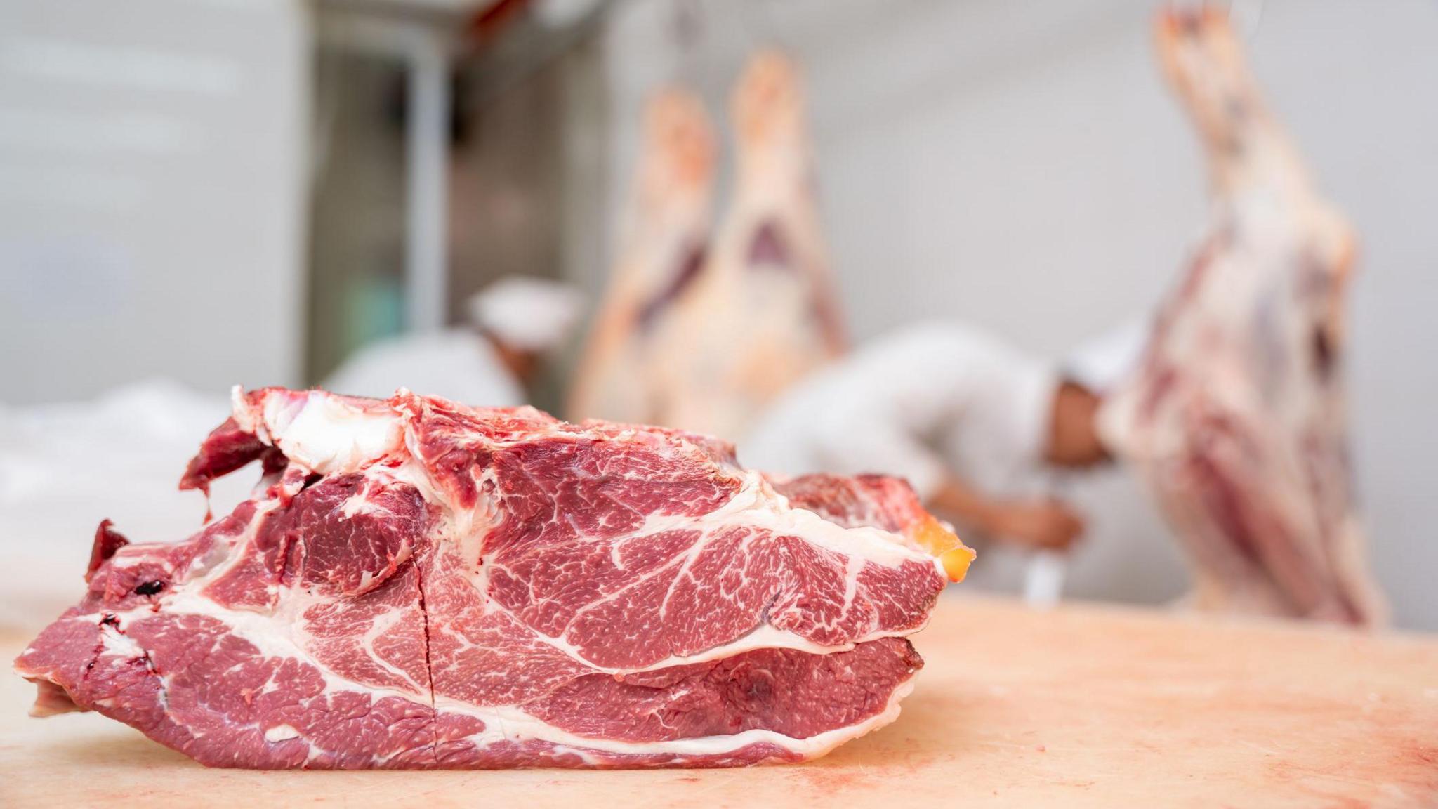 A large slab of meat on a wooden counter while workers look busy in the background