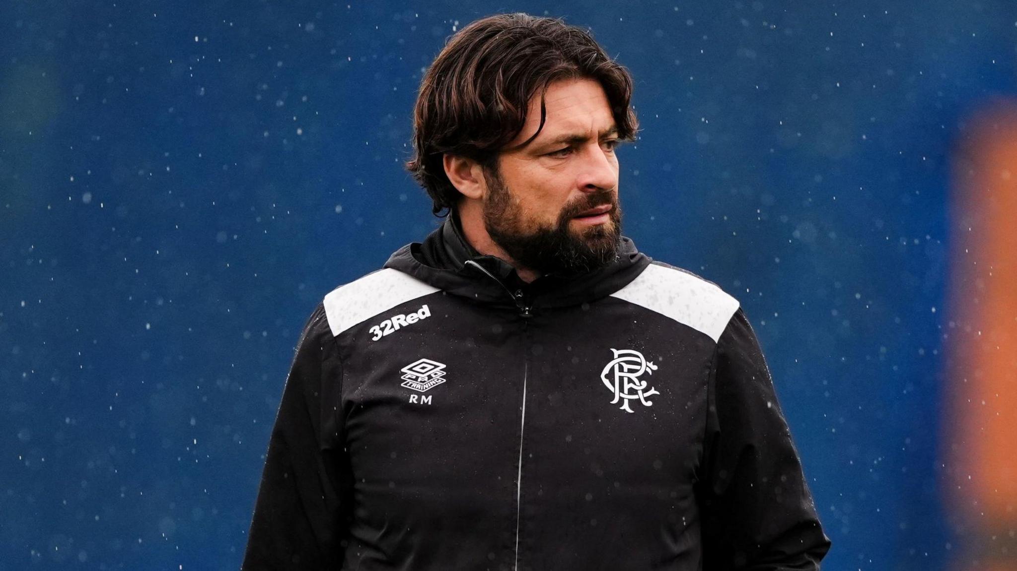 Russell Martin looks off to his left. He has black hair, combed in a middle parting, and a beard. He is wearing a black jacket with the Rangers crest and Umbro logo on it. He has been photographed in the rain against a blue backdrop.