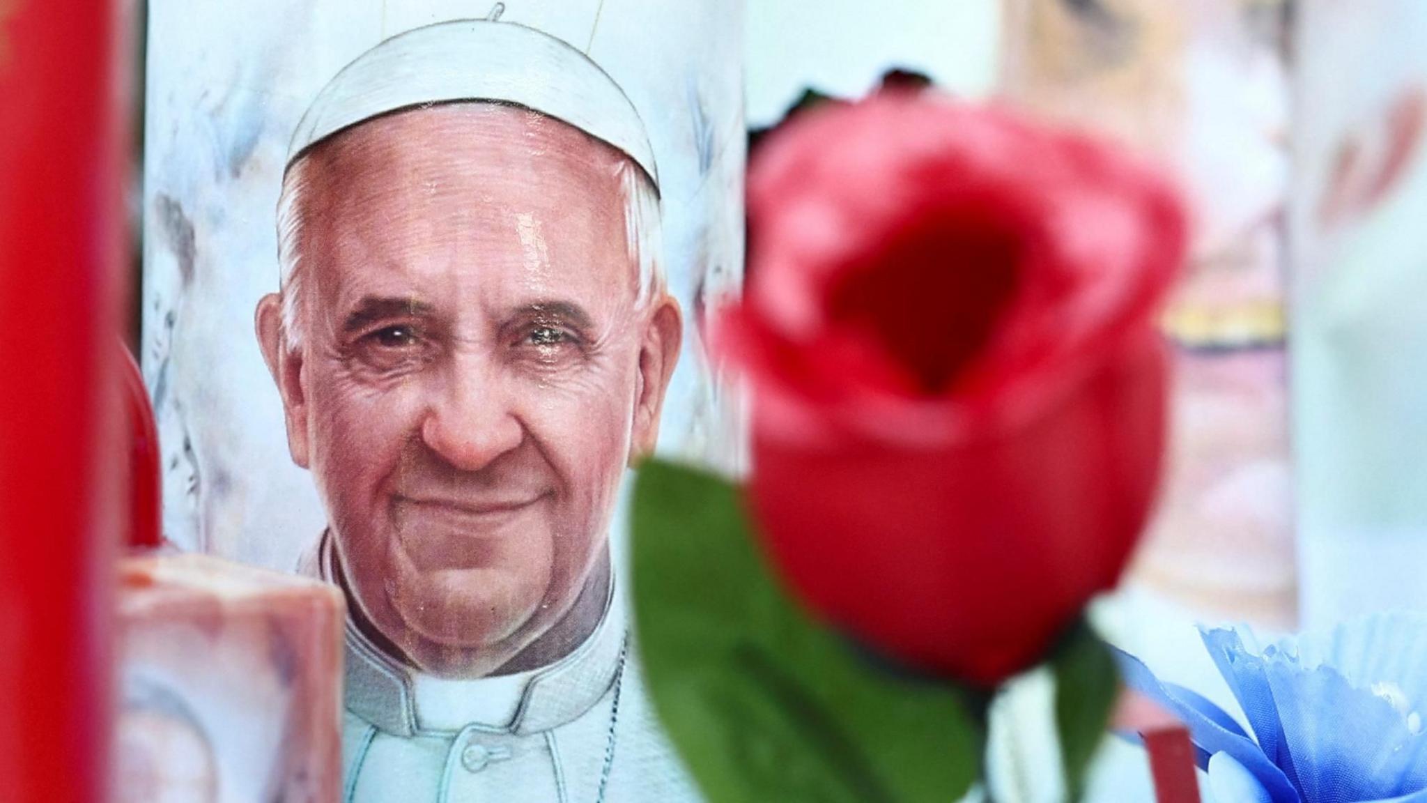 On the left, a lifelike painting of the Pope on a cylindrical surface, likely a candle, and on the right, a red rose 