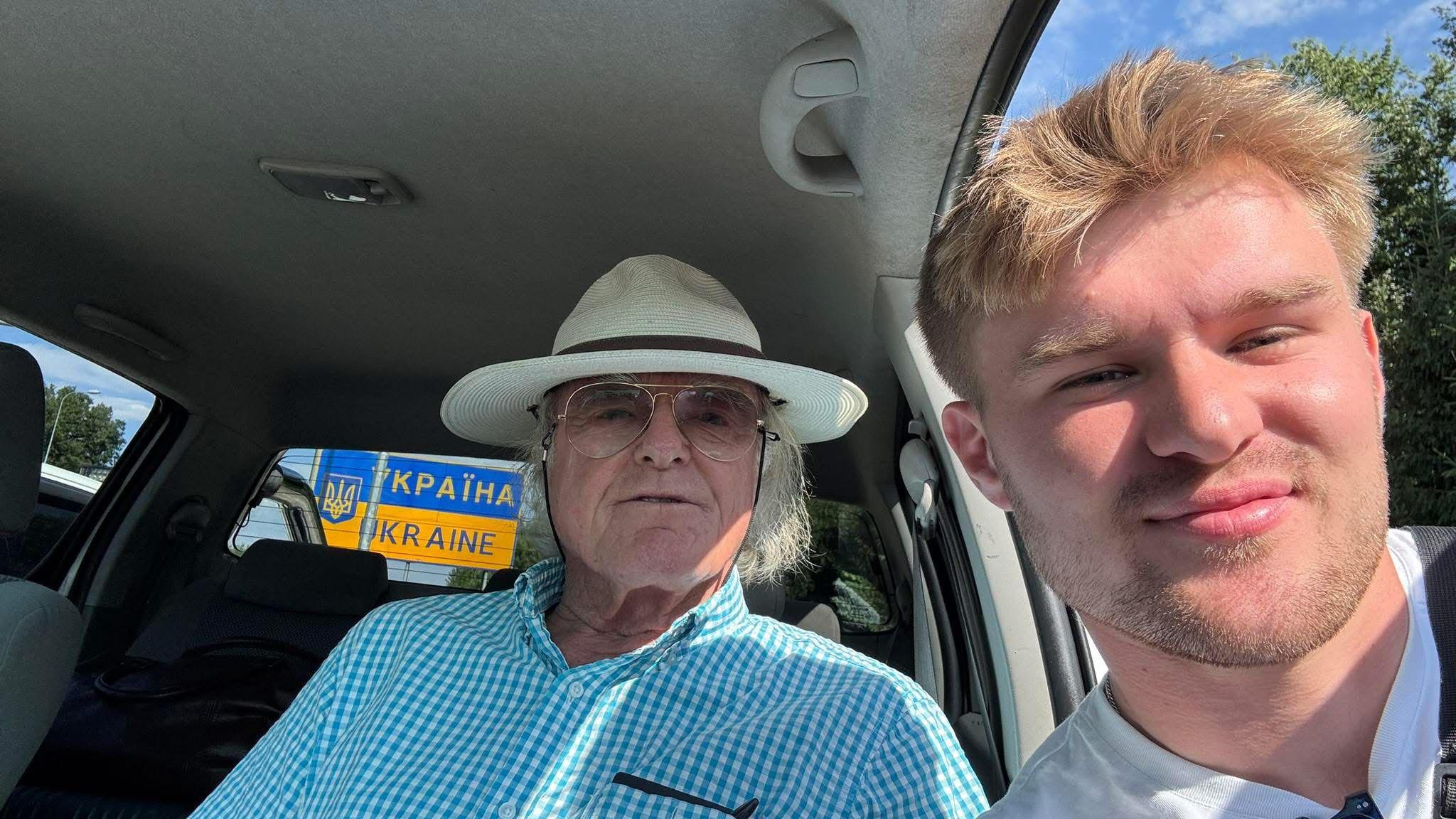 John Caldwell, sat in a vehicle, is wearing a white hat and Fynn Watt, is leaning through the vehicle's left front window, as they both pose for the picture with a sign for Ukraine visible through the vehicle's back windscreen.