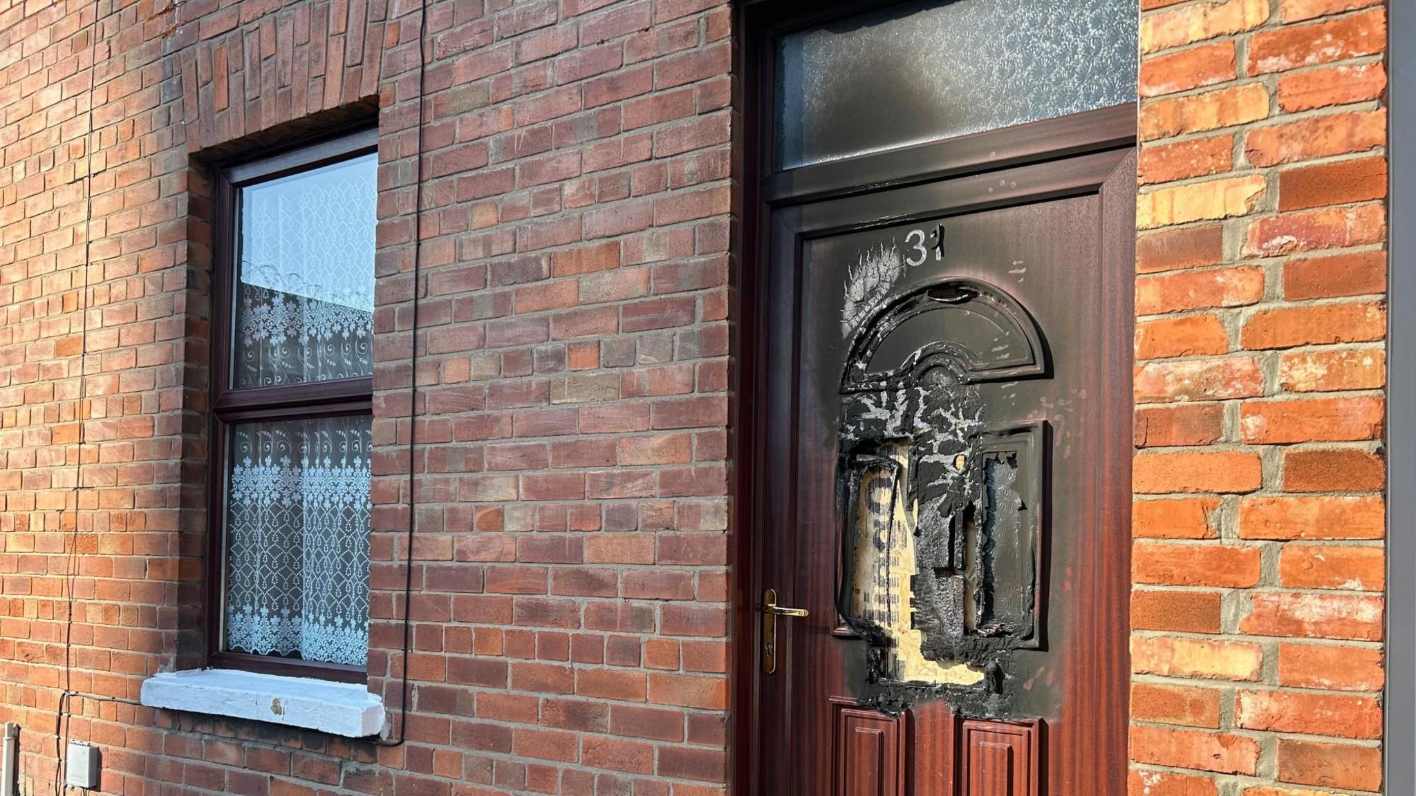 A red brick house with a burn mark on the front door. The door was originally brown but has a large black mark on over half of the door, there is also a hole in it. The window seems to be untouched