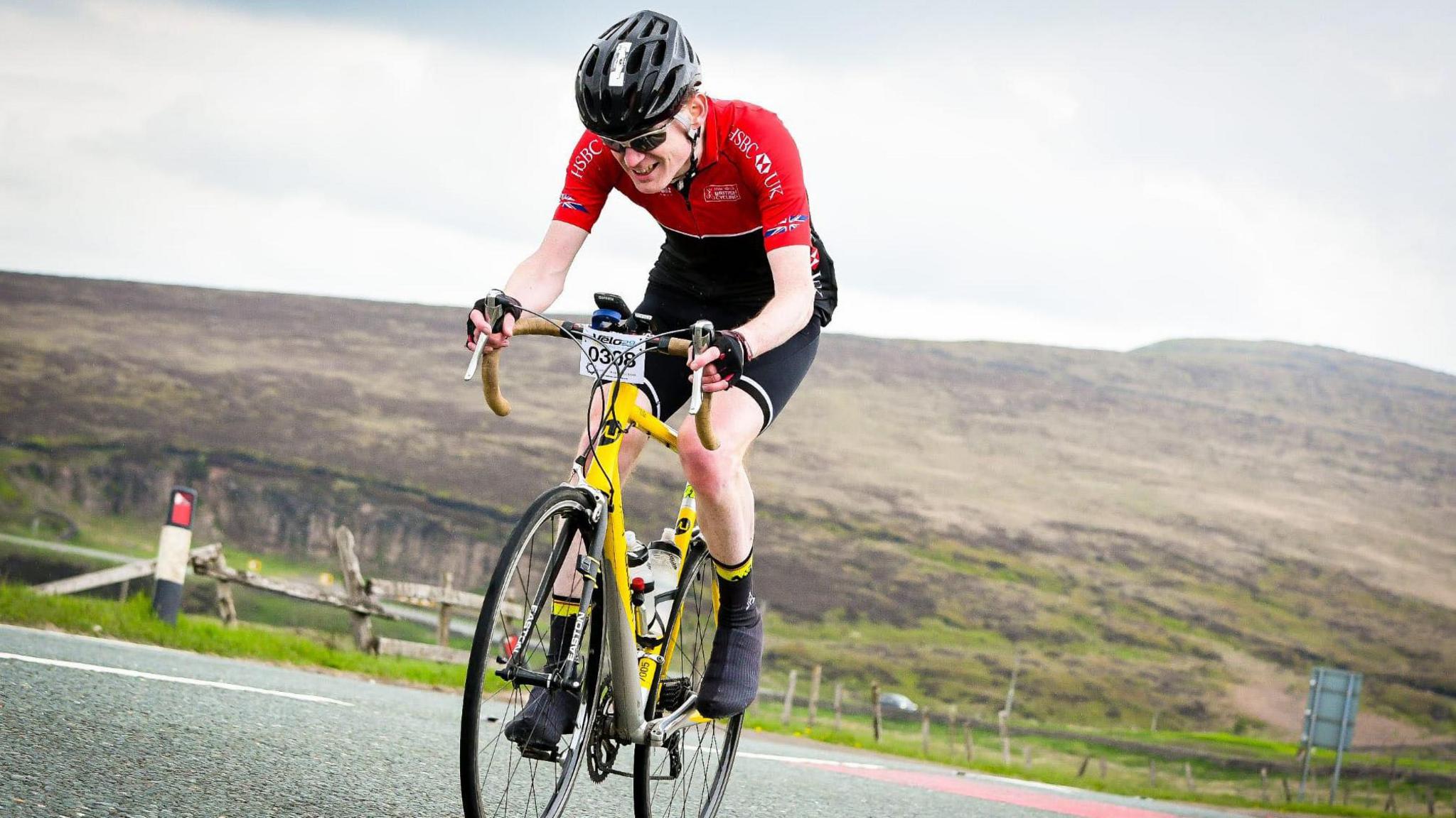 A road cyclist wearing a red jersey rides a yellow bike through a mountainous route.