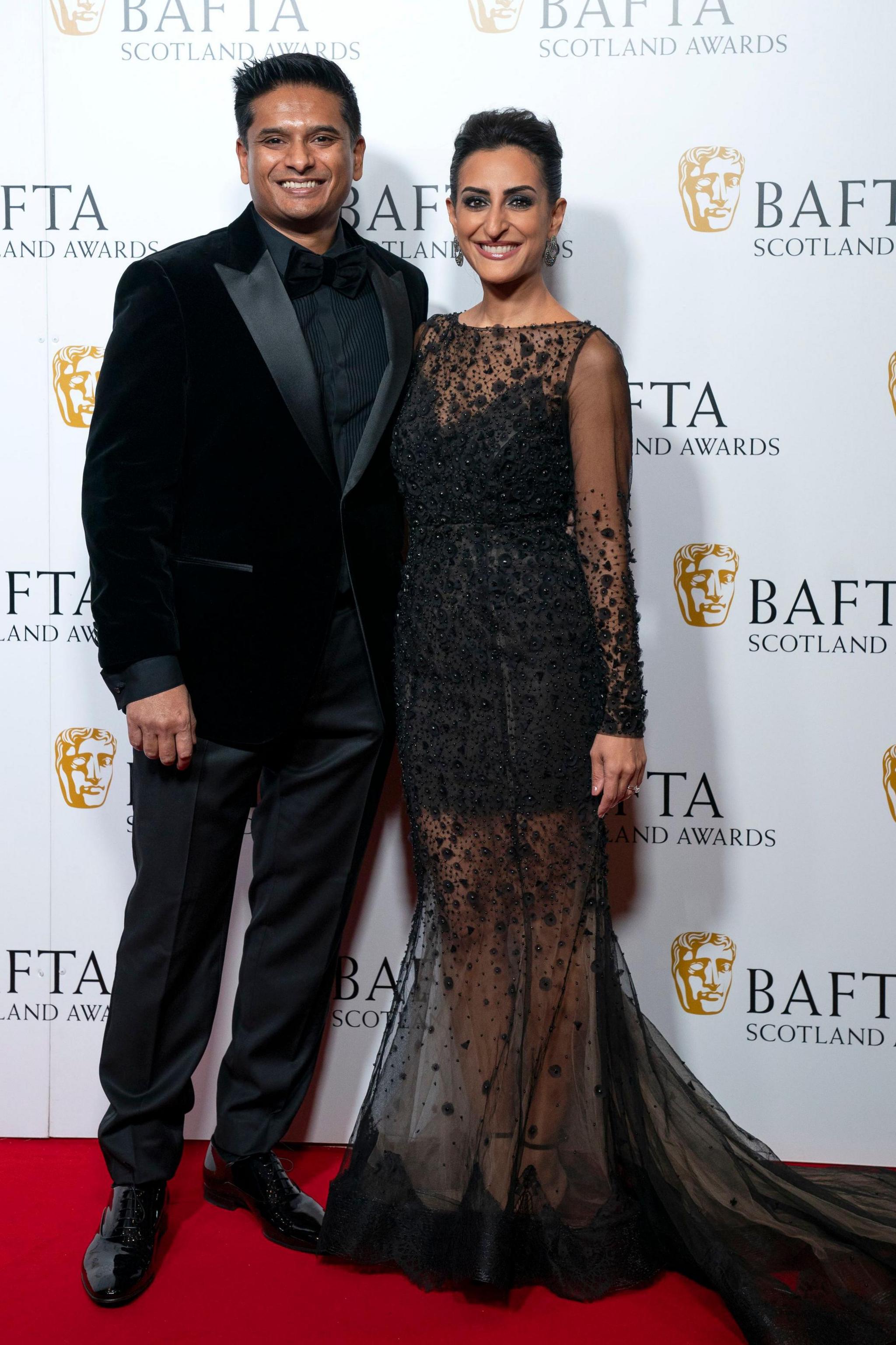 Dr Sandesh Gulhane, in a black suit, and Dr Punam Krishan, in a long black dress with lace train, stand in front of a Bafta Scotland backdrop