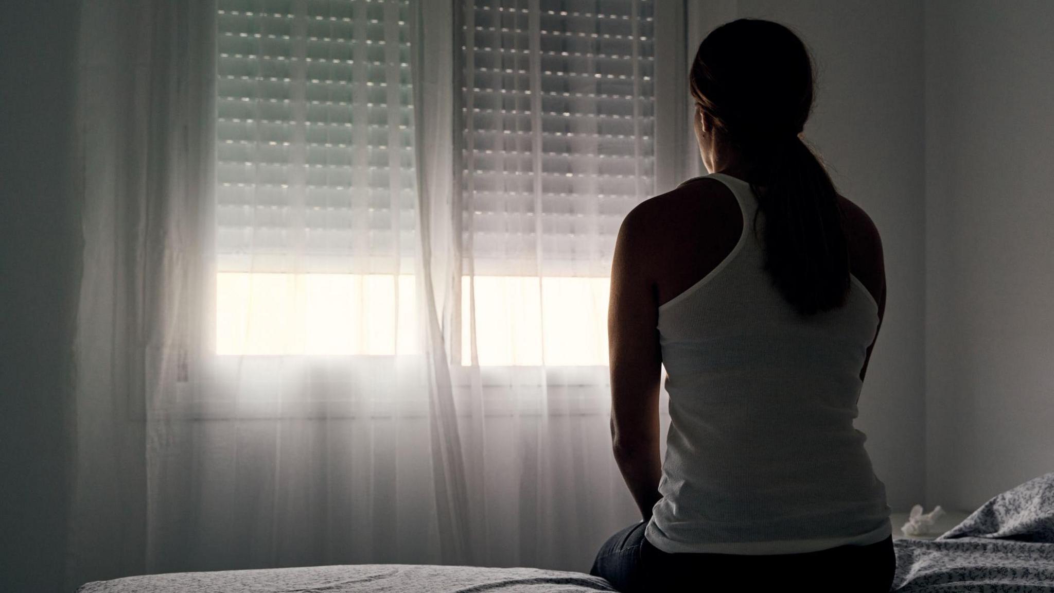 A still of a woman sitting in a darkened room with her back to the camera. She is sitting on a bed wearing a white vest tshirt staring out of a window which has the blinds pulled almost to the bottom and sheer curtains pulled across.