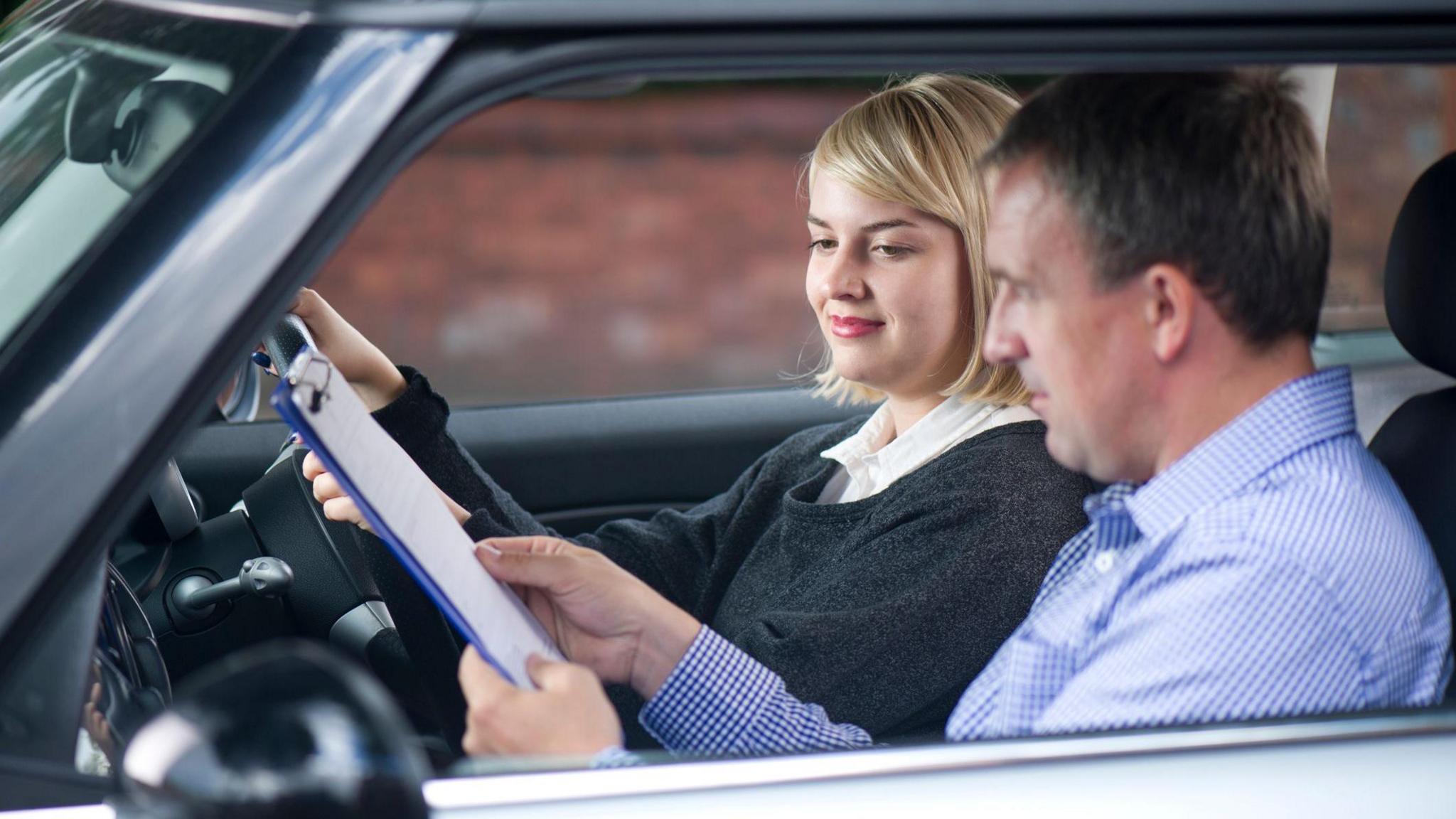 A young woman taking a driving test. She has blonde hair and is wearing a dark grey jumper. Her tester is a man in a check blue shirt. He is showing her a document on a clipboard.