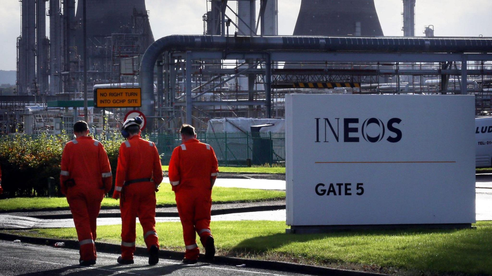 Three workers in orange boiler suits walk past a white Ineos sign. The Grangemouth plant can be seen in the background.