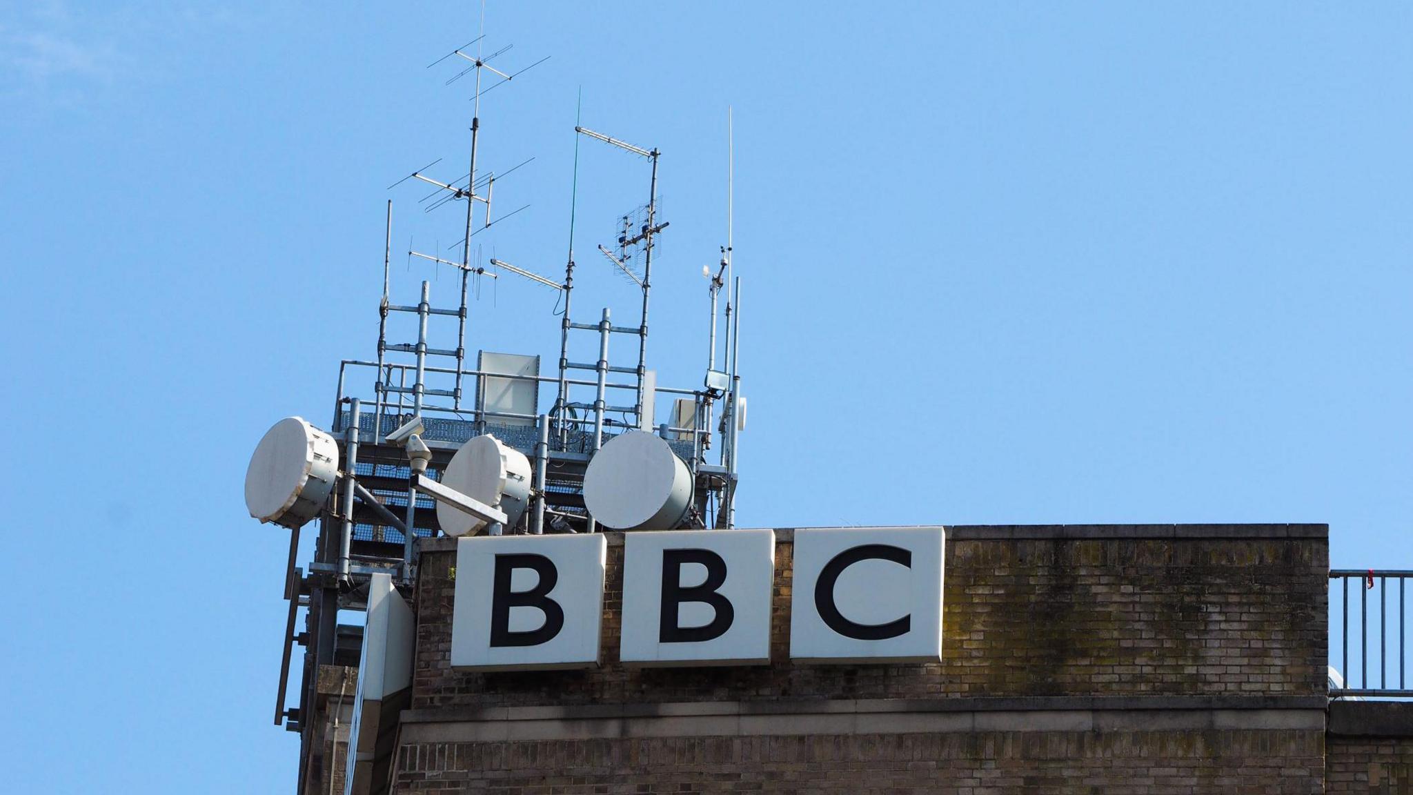 antennae and satellite transmitters against a blue sky, on top of a roof where a sign reads BBC in very large letters