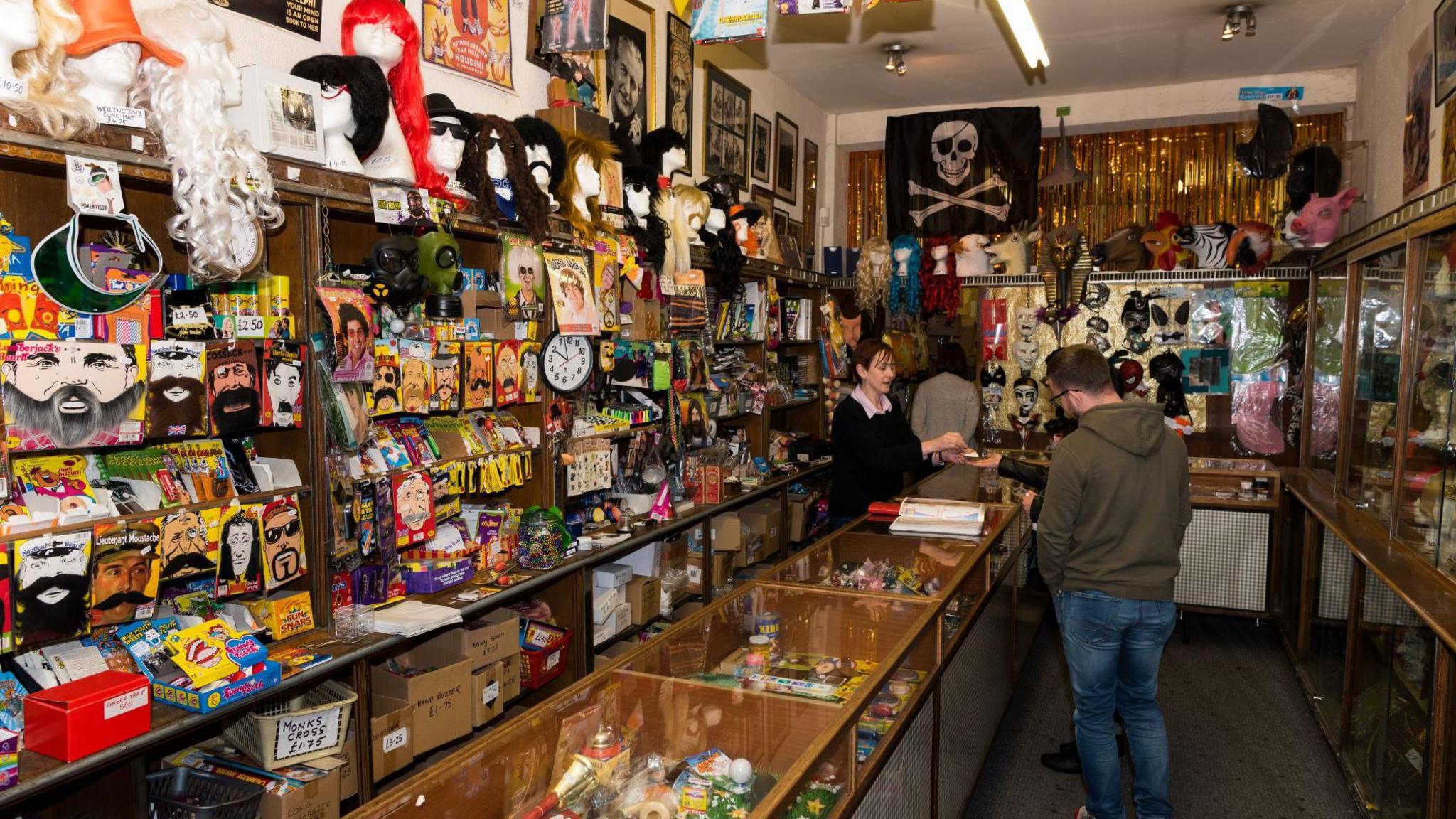 Interior of Tam Shepherds, where a female staff member is serving customers. It is a small rectangular shop with a glass-topped counter, walls covered in costume props, joke items and gag gifts.