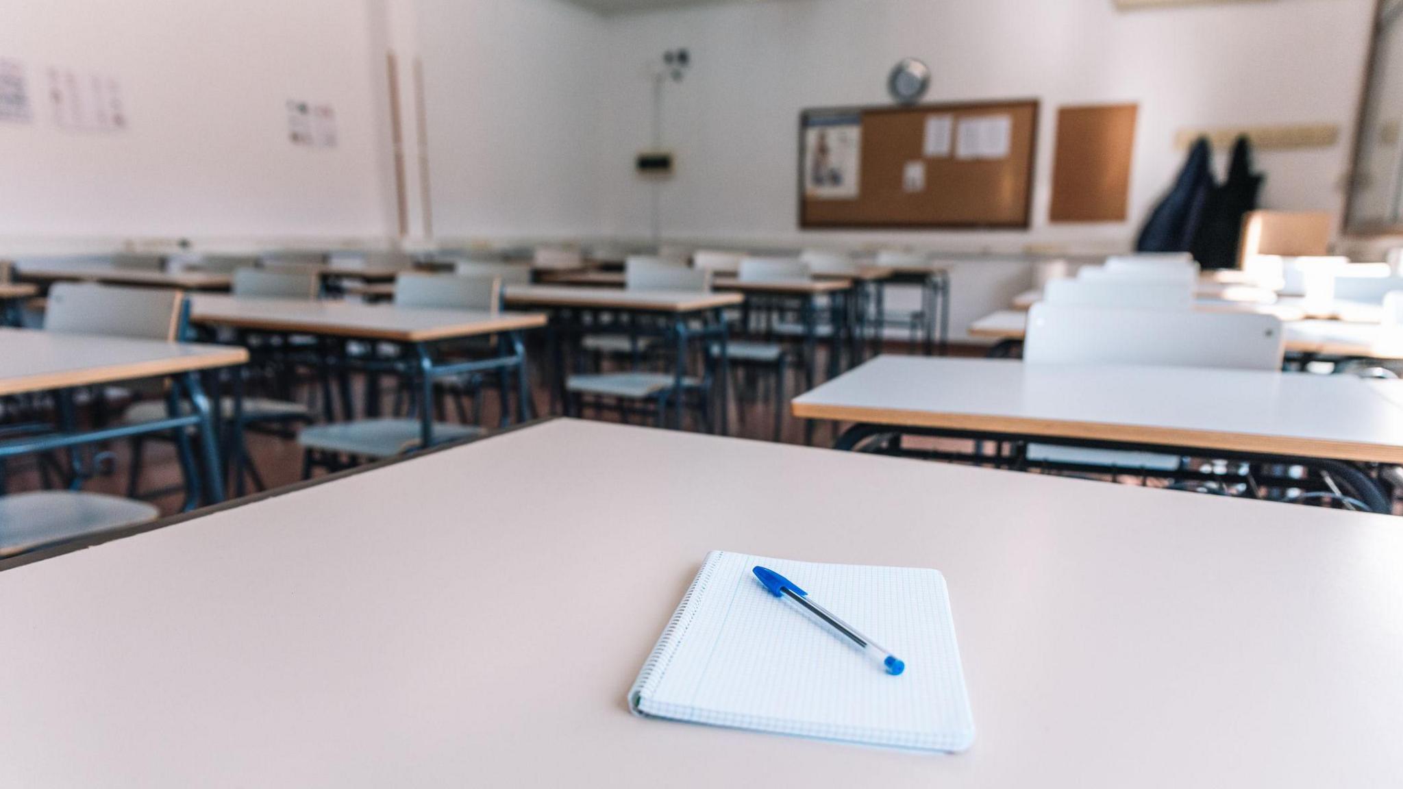 An empty classroom, with desks and chairs, and a pad of paper and a pen on the closest table to the camera 