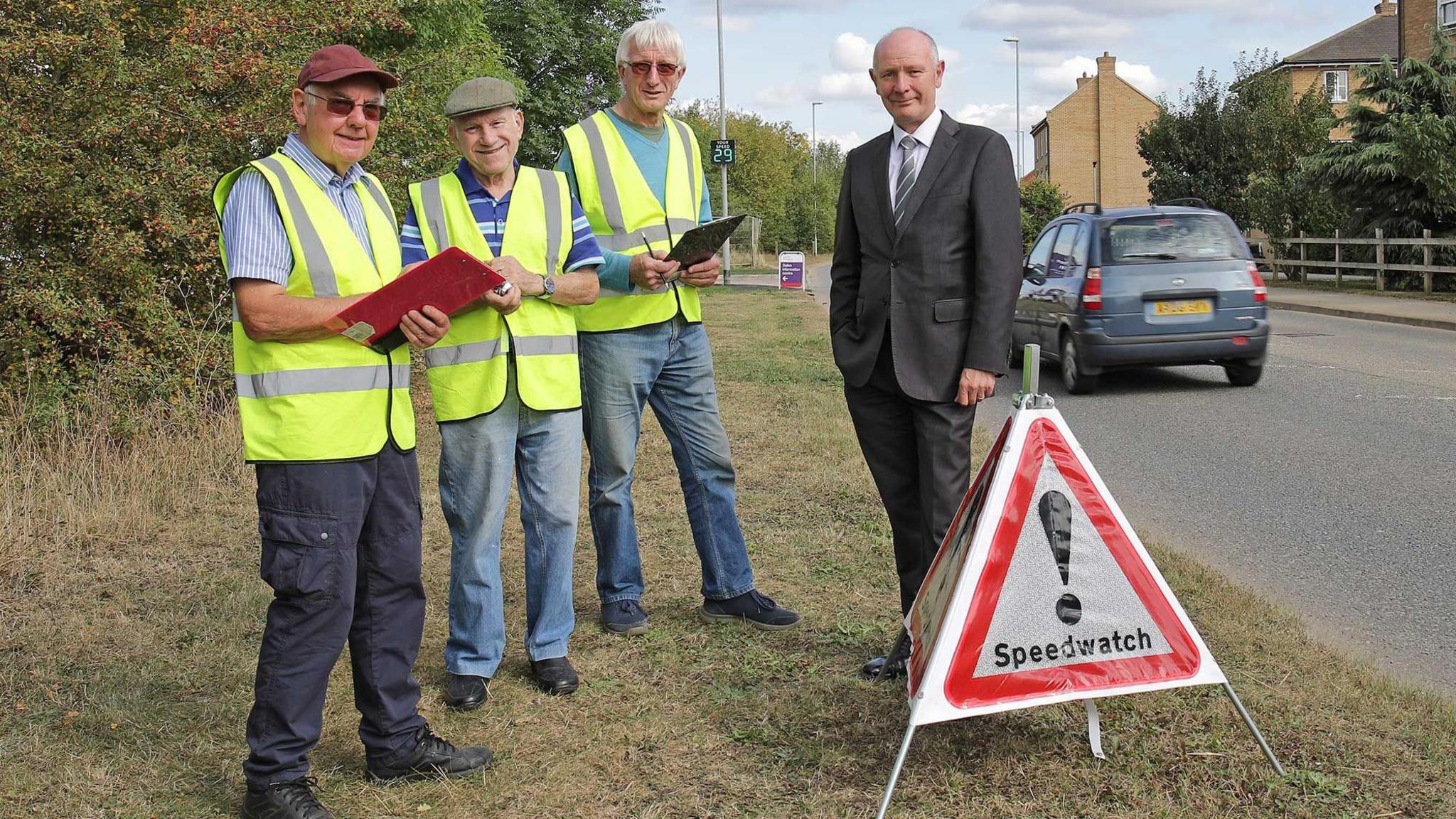 Three men in hi-vis jackets, two of them holding clipboards, are standing next to Darryl Preston wearing a suit and striped tie. In front of them is a triangle with Speedwatch written on it, and an exclamation mark.