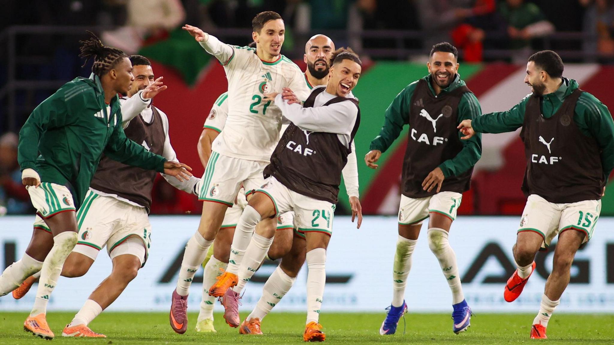 Adil Boulbina, wearing a white Algeria kit bearing the number 27 on the chest, is chased by seven joyful team-mates after scoring a goal