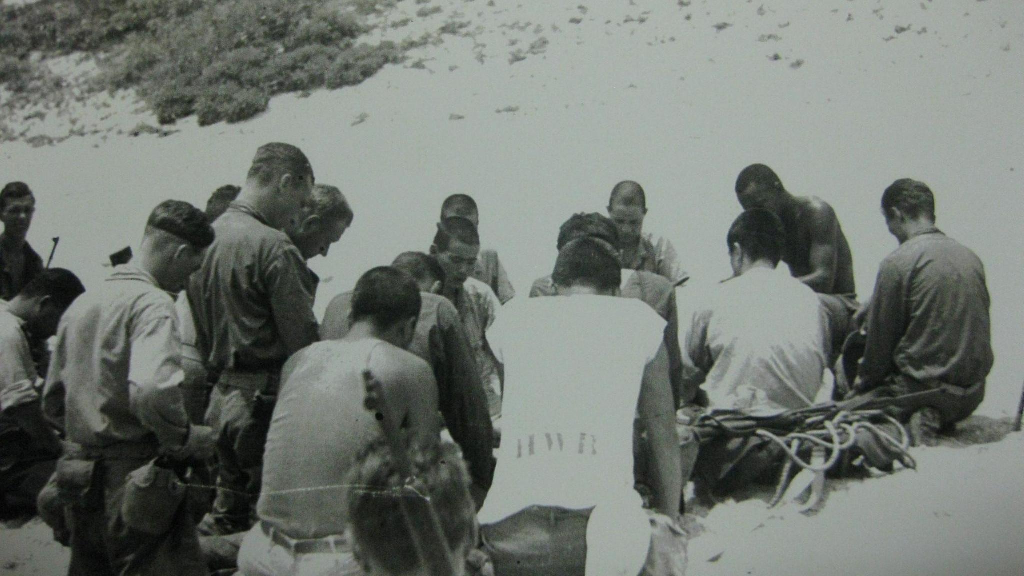 A black and white photograph shows a group of about 20 men sitting and kneeling in prayer on a beach. Some of the men are Japanese, some are American. They all have short hair and have their heads bowed.