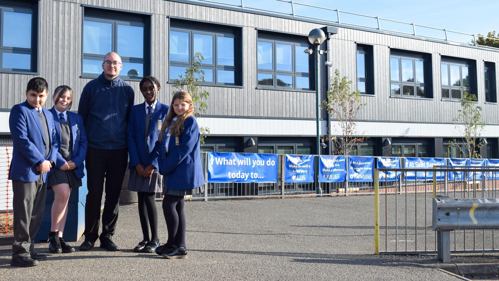 Head of year 7 Adam Tomlinson stood with four pupils in front of a building with grey panels  and windows with black frames