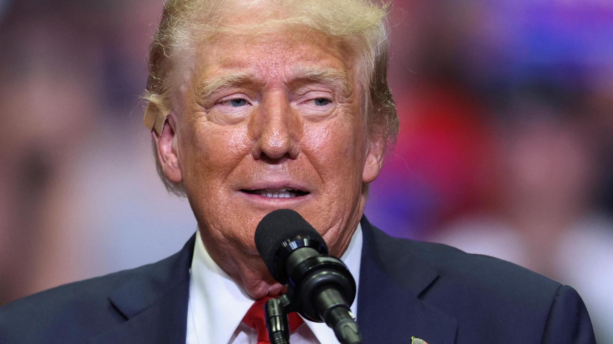 Donald Trump speaking into a microphone during a political rally. the blurred crowd can be seen behind him.