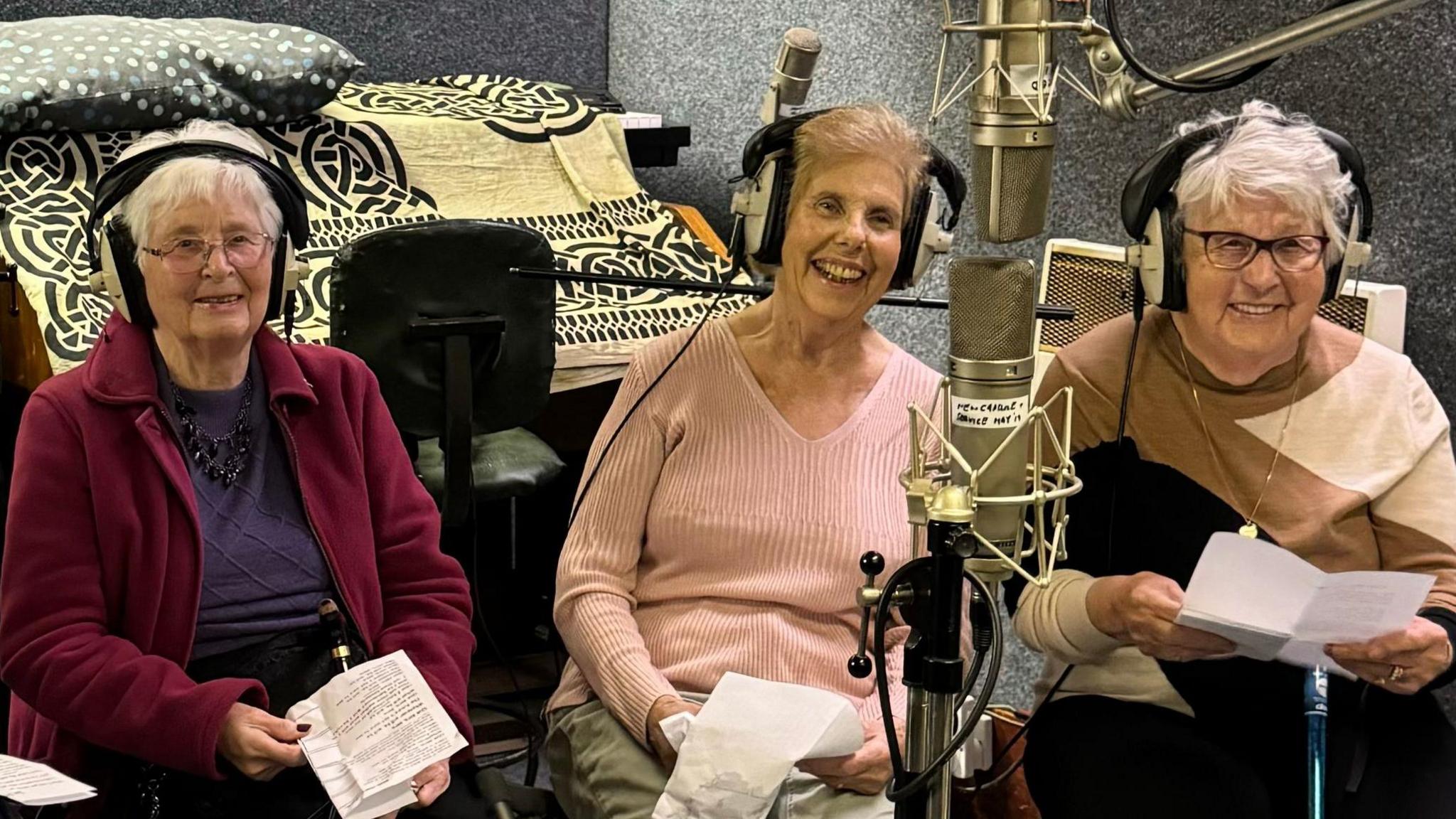 Three elderly women, seated and wearing headphones, smile as they prepare to sing into large, metallic microphones in a recording studio. Behind them is an upright piano, covered in rugs, and soundproofed grey walls.