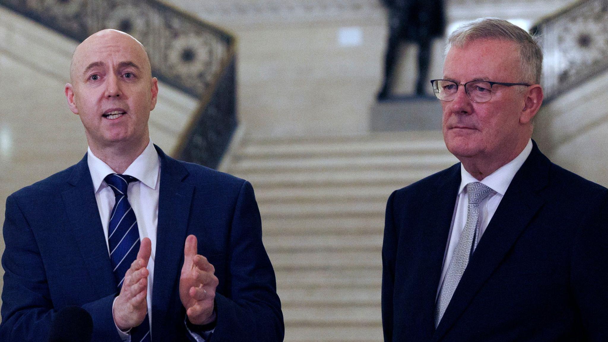 Jon Burrown and Mike Nesbitt. Jon os bald, wearing a white shirt and blue suit. Mike has short grey hair, wearing a white shirt, grey tie and black blazer.