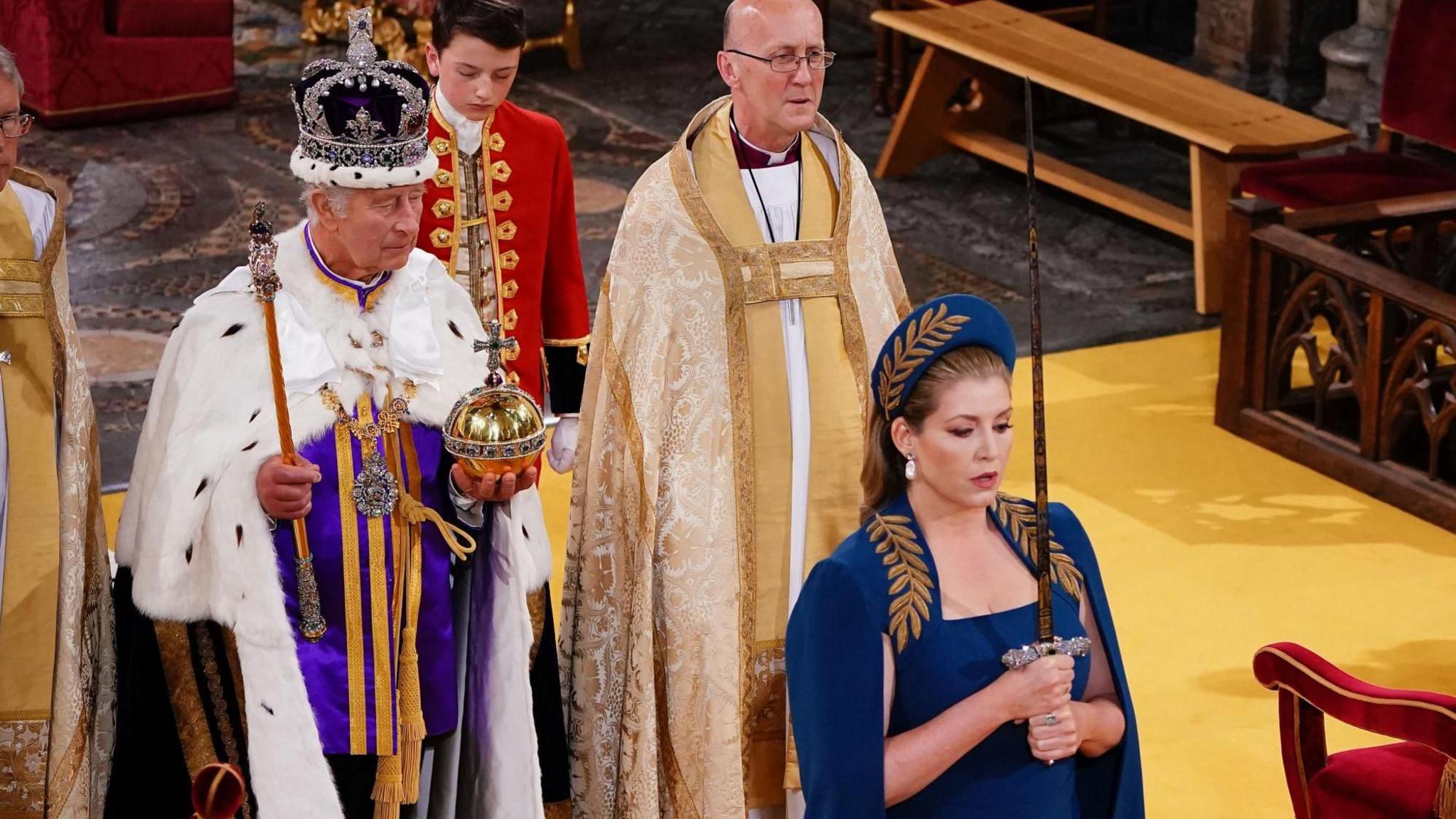 A newly-crowned King Charles III stands holding an orb and sceptre, while Penny Mordaunt, wearing a blue dress and hat, stands in front of him holding a sword with both hands. The sword is held upright in front of her face.