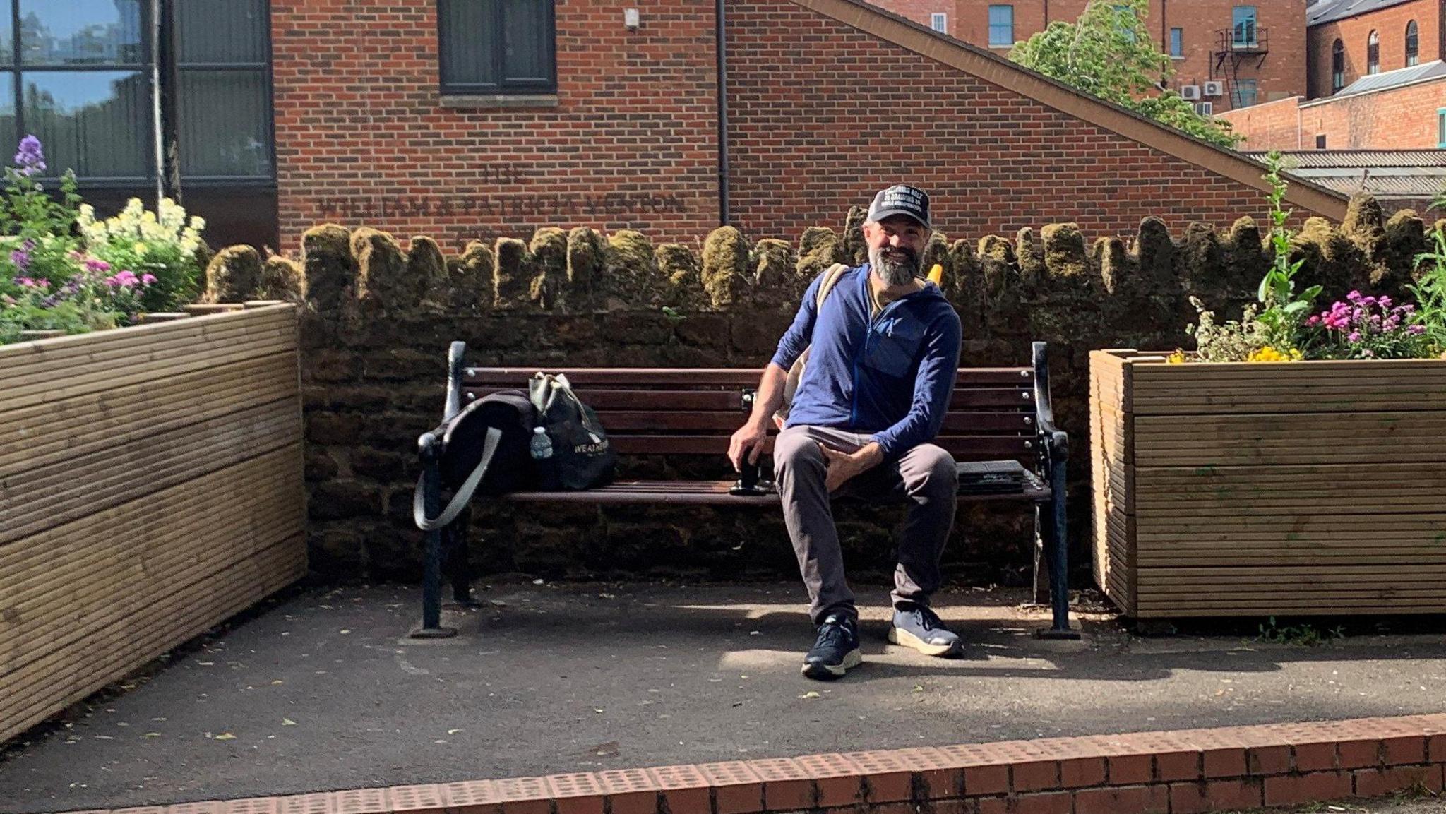 A man sitting on a wooden bench, smiling, looking at the camera and wearing a blue top, trousers, trainers and a cap. He has two large flower planters beside him.