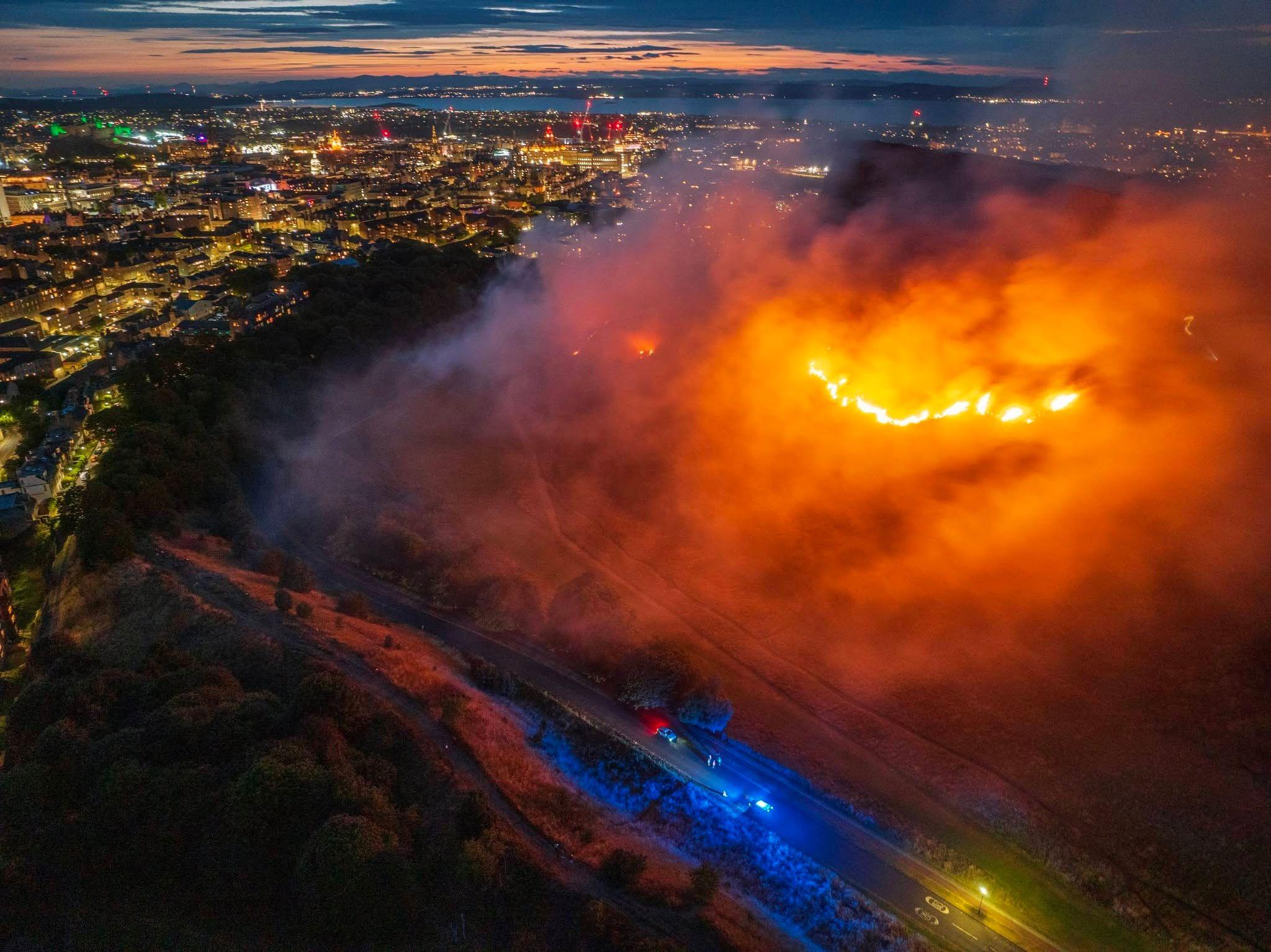 A line of fire on a hillside with city lights behind and blue lights of emergency vehicles in the foreground