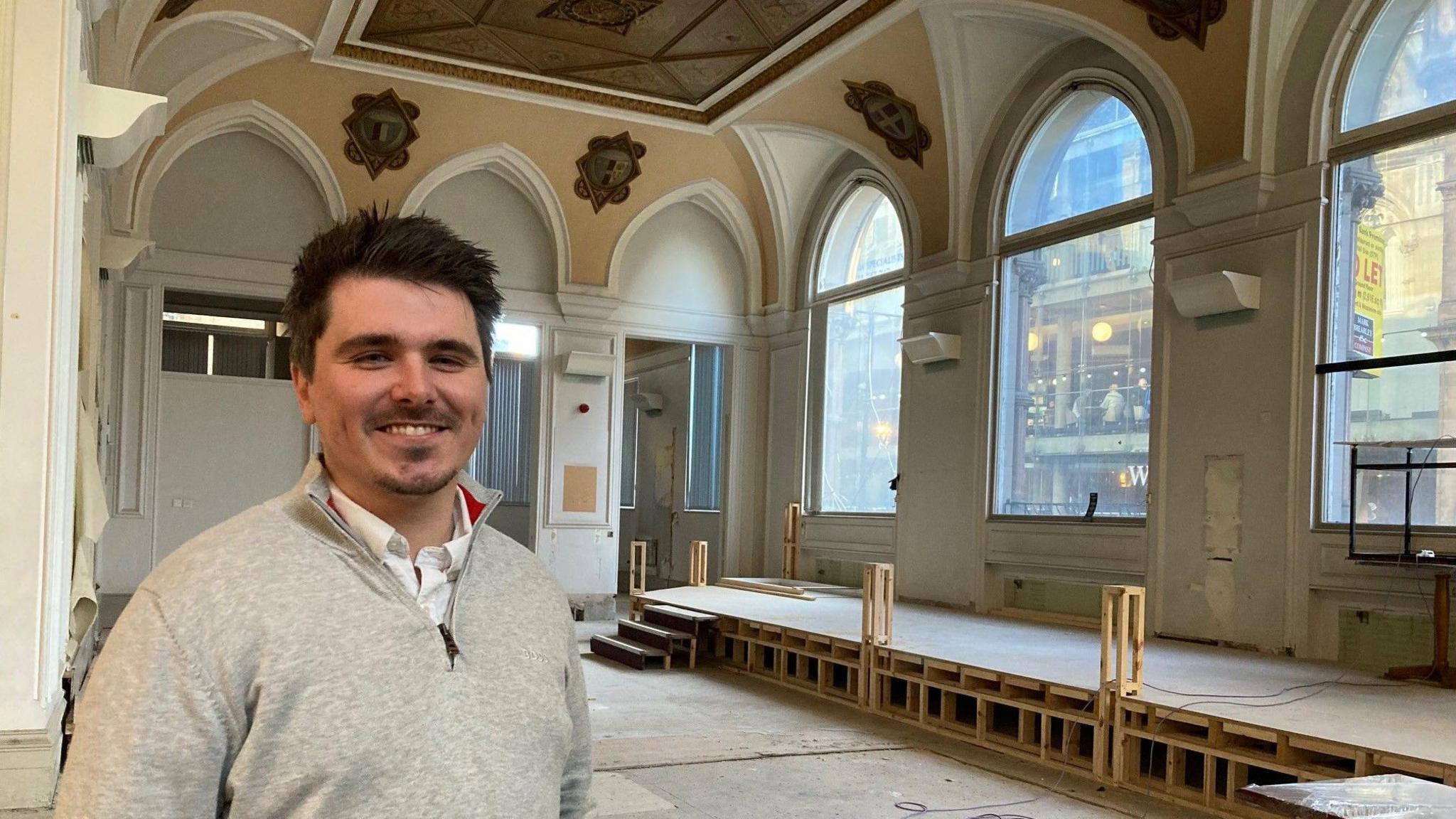 A man with dark hair, a goatee and wearing a grey zip-up top in a striped out room with high ceiling and three large, arched windows.