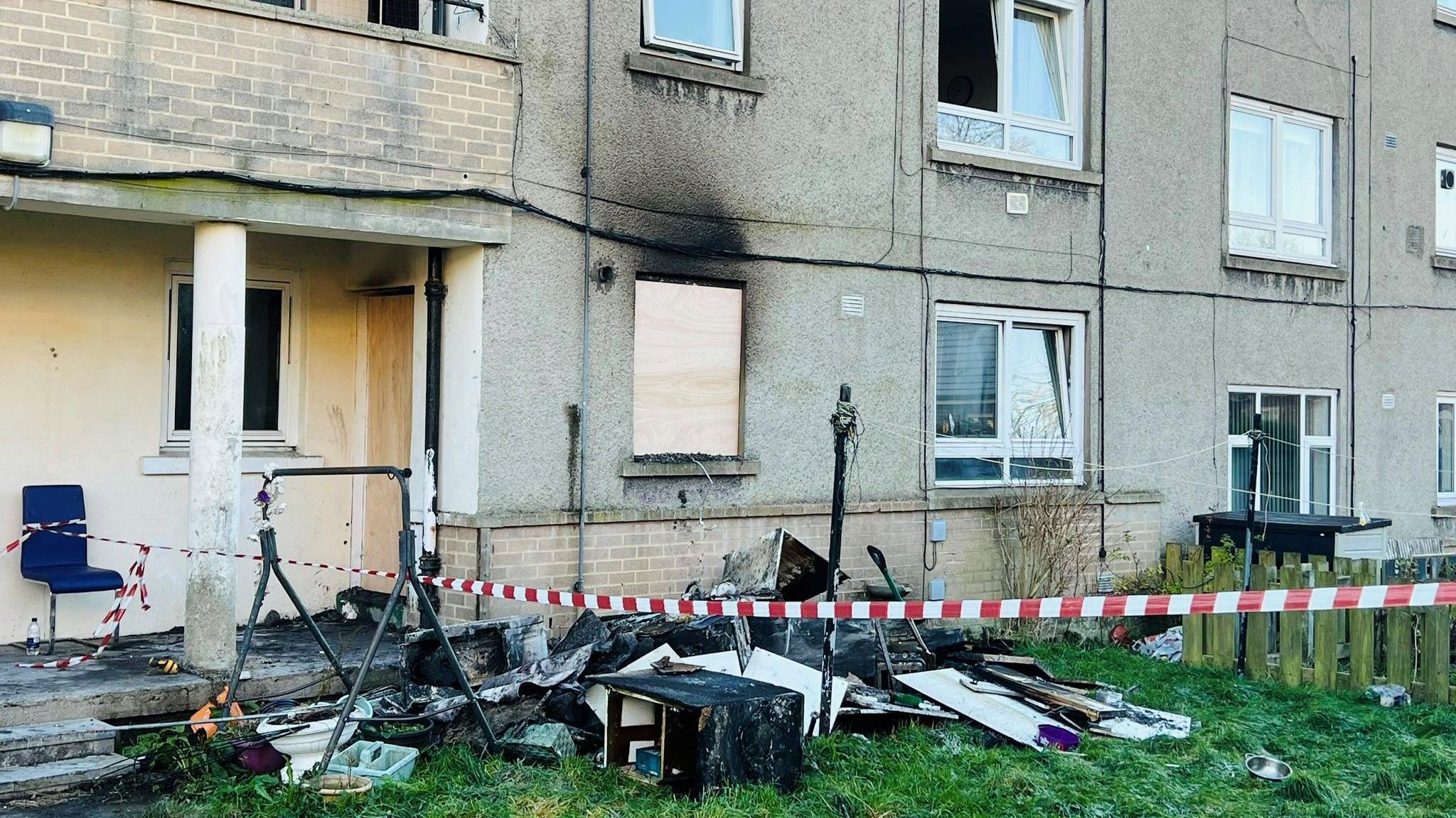 Image of flats after fire, charred walls and damaged items on grass, with red and white taping.