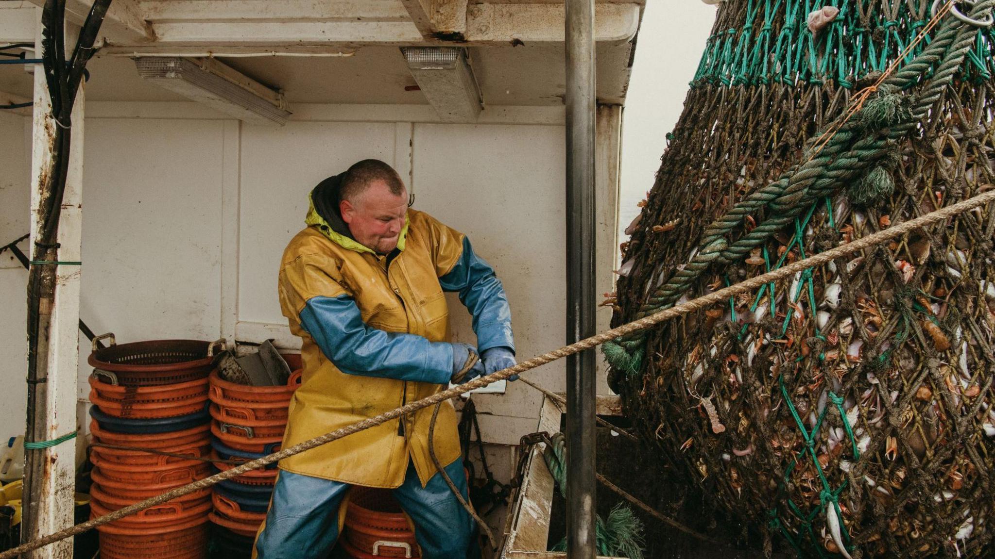A colour image of a fisherman hauling in a large net full of fish. He is wearing yellow and blue waterproofs and using his full strength to haul in the net. His face is straining. 