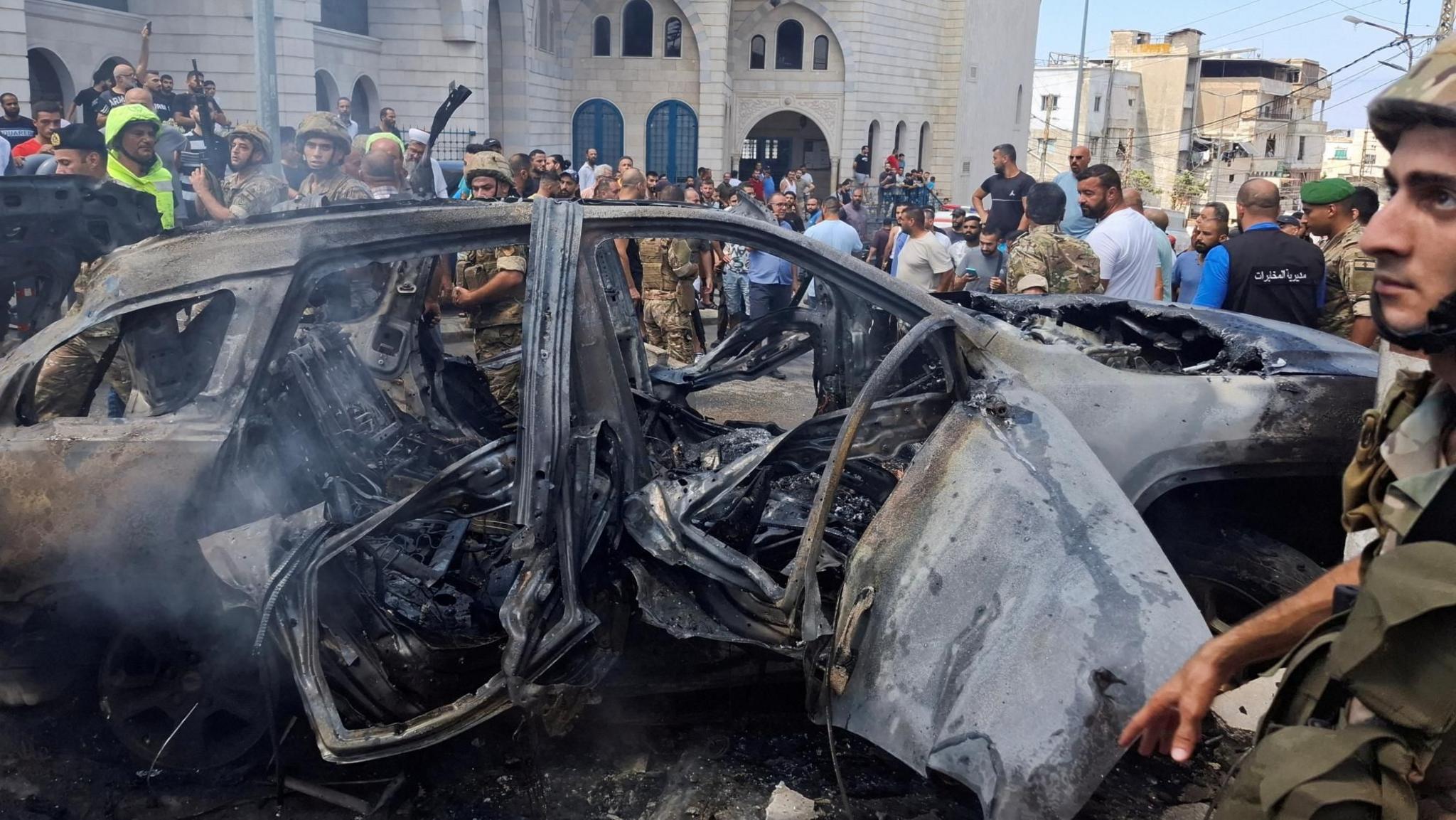 People stand near a burnt car after an Israeli strike on the outskirts of the southern Lebanese city of Sidon (21 August 2024)