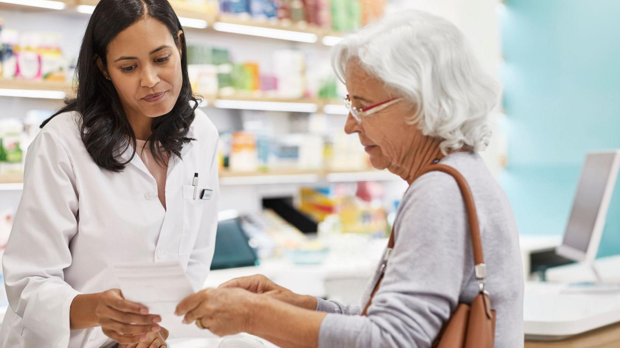 An older woman with grey hair is asking a pharmacist, who has long dark hair and is wearing a white coat, about her prescription in a chemist shop