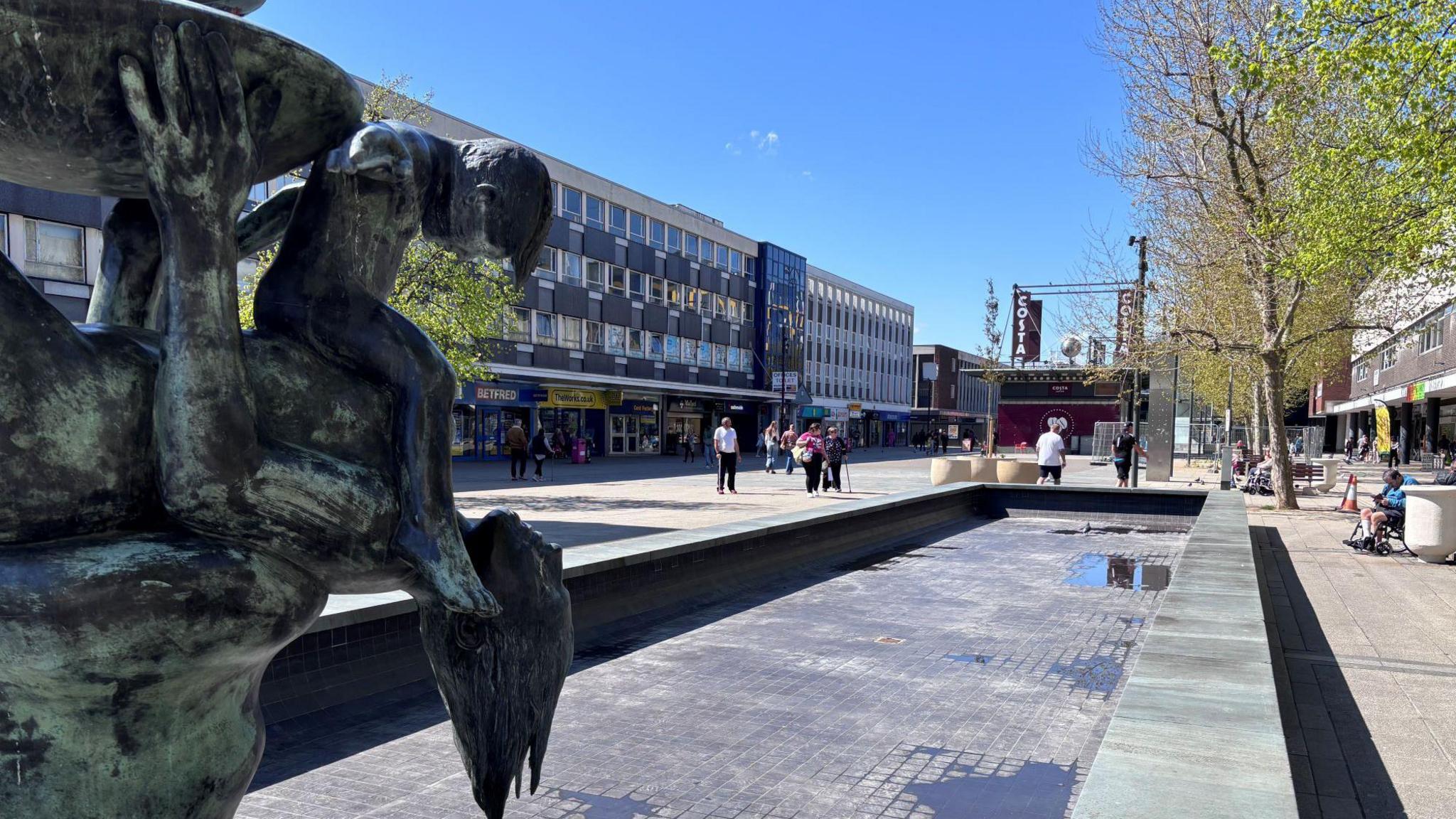 A pedestrianised town centre street featuring shops, a statue and an empty water feature.