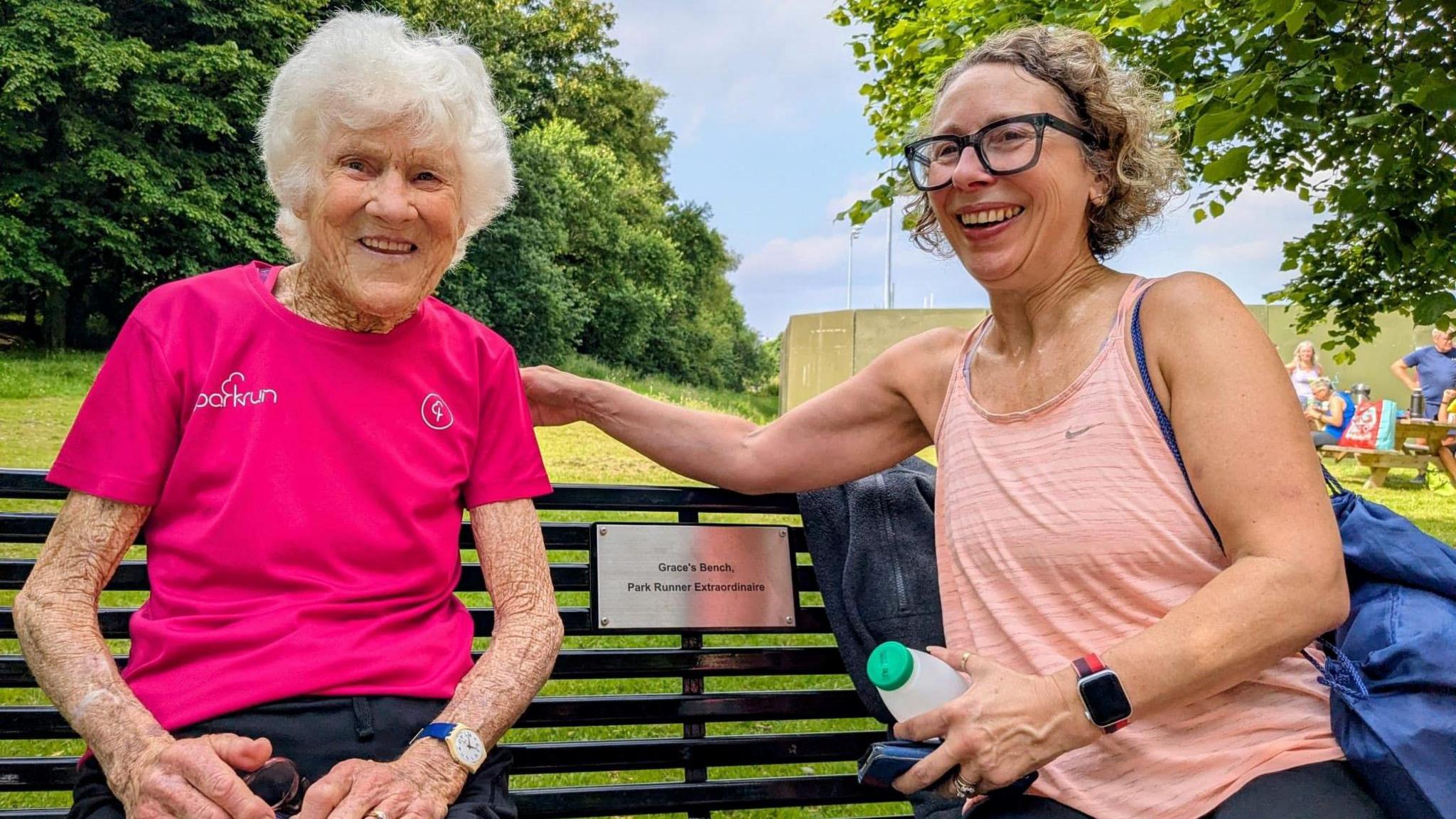 Grace Chambers wearing a pink parkrun t-shirt. She is looking at the camera, She is sitting on a black park bench, there is a small plaque on the bench which says Grace's bench - Park Runner extraordinaire. She is sitting with another woman in a peach colour running top and black glasses