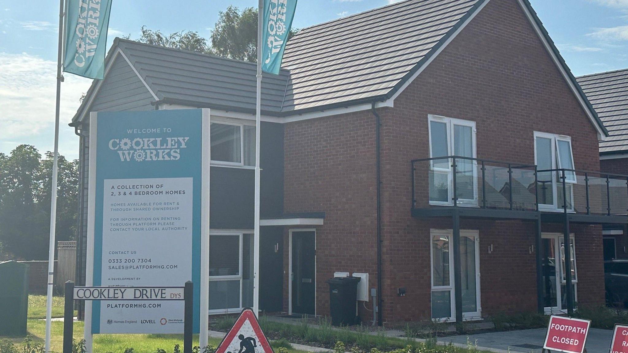 Road entrance, with signing welcoming people to Cookley Works, in front of a new house and 'footpath closed' sign 